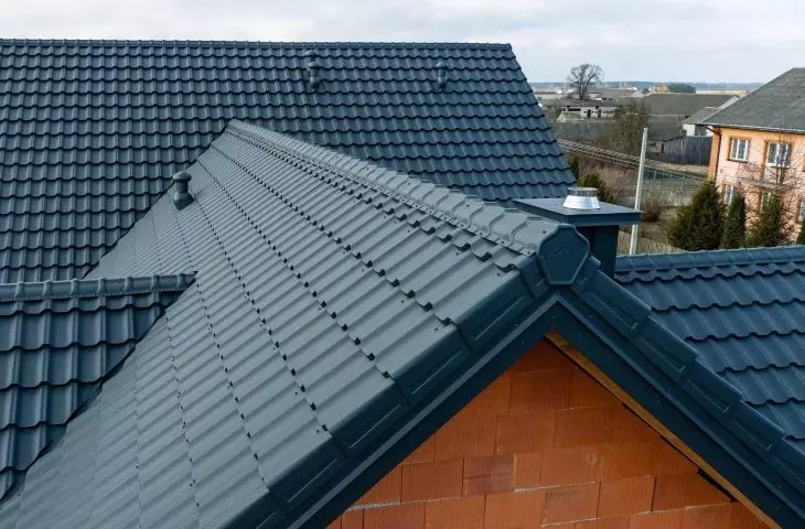 Nowoczesny, grafitowy dach z dachówki ceramicznej, widok z góry na fragment komina z metalowym zakończeniem. W tle zabudowa jednorodzinna i drzewa.