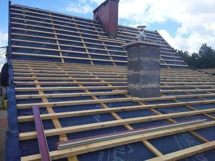 Dach w trakcie krycia: drewniana konstrukcja łat na czarnej membranie, komin z szarej cegły. Widoczne elementy blacharskie. Błękitne niebo w tle.