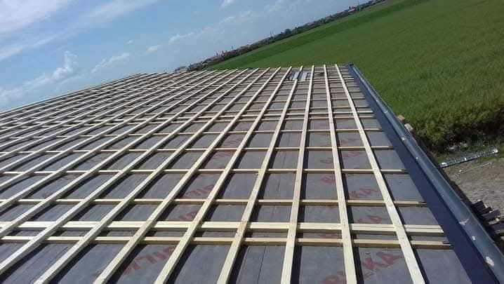 Więźba dachowa z łatami na folii w trakcie montażu, widok z góry na tle zielonego pola i błękitnego nieba. Elementy konstrukcyjne dachu w regularnym układzie.