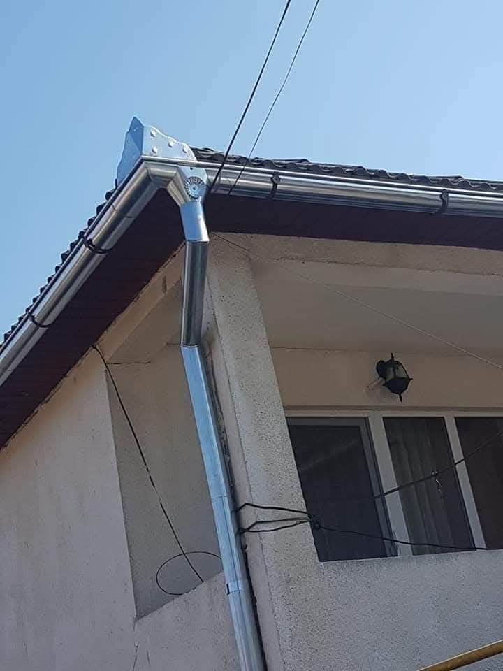 Nowa, błyszcząca, srebrna rynna pionowa i pozioma, zamontowana na budynku z brązowym dachem. Widok z dołu, błękitne niebo w tle.