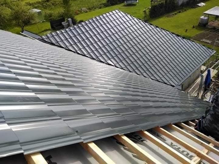 Montaż blachy trapezowej na dachu domu jednorodzinnego. Widoczna więźba dachowa i częściowo ułożone pokrycie w kolorze szarym. Ujęcie z góry, w tle zieleń.