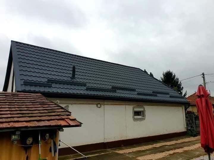 Grafitowy dach z blachy trapezowej na parterowym budynku z białą elewacją, widoczny z boku, z fragmentem dachu sąsiedniego budynku i czerwonym parasolem w tle.