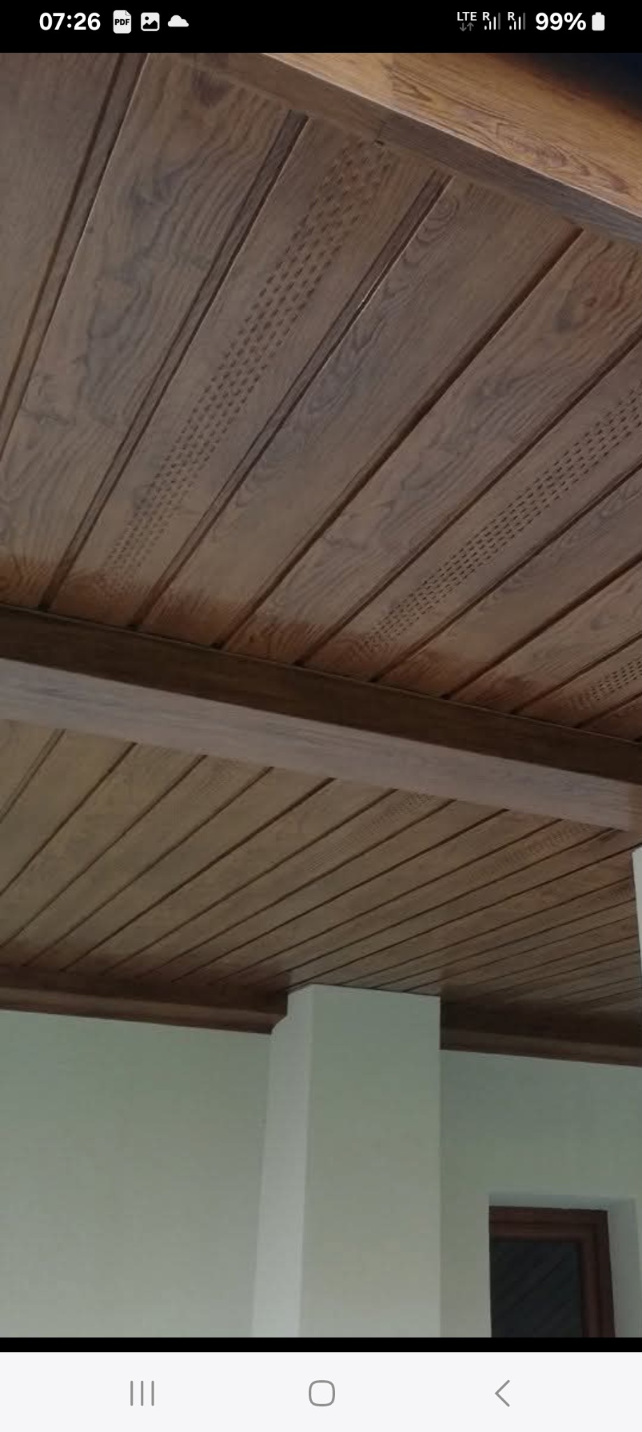 Drewniany sufit z dekoracyjnymi żłobieniami i listwami, widok pod kątem. Jasne drewno, regularny wzór, fragment ściany w tle.