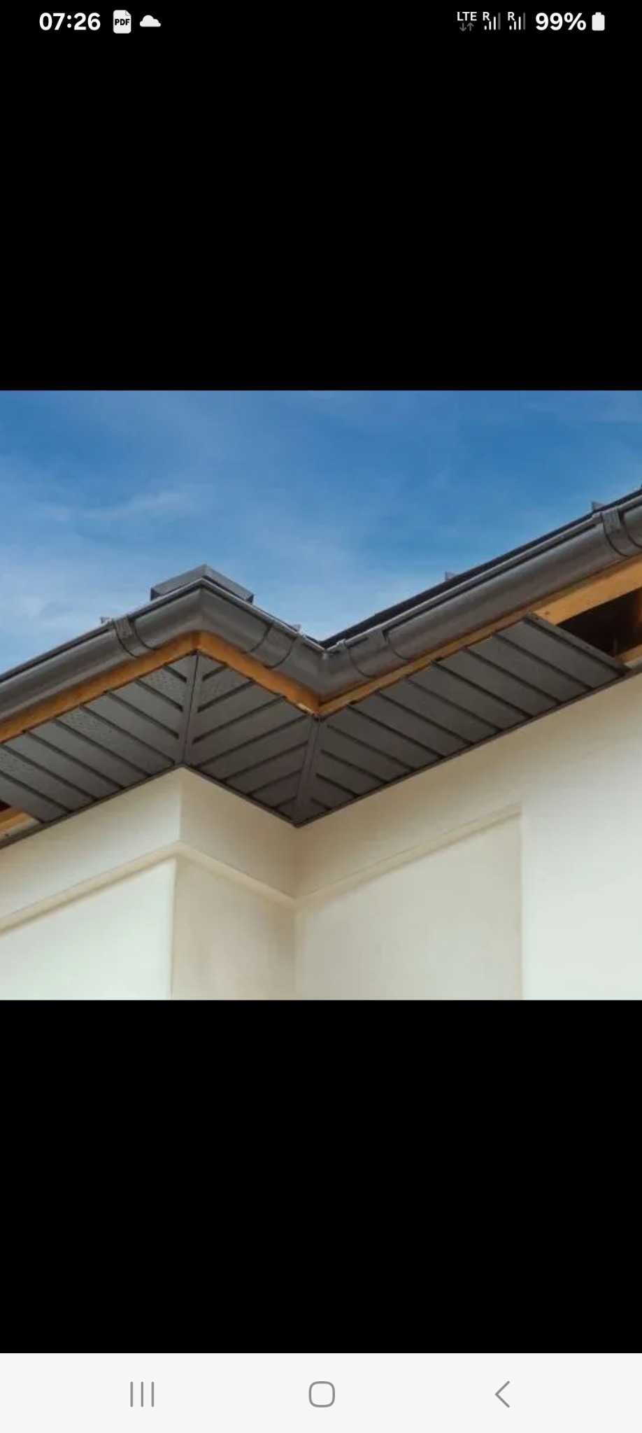 Szczegółowe ujęcie narożnika dachu z ciemną blachą trapezową, widoczne rynny i drewniane elementy konstrukcyjne na tle błękitnego nieba. Eleganckie wykończenie.