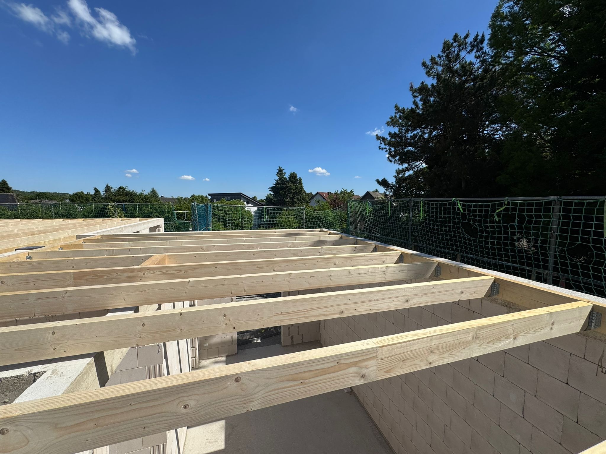 Wiązar dachowy z jasnego drewna na murze z bloczków, widok z dołu w słoneczny dzień. Elementy konstrukcyjne dachu, błękitne niebo w tle.