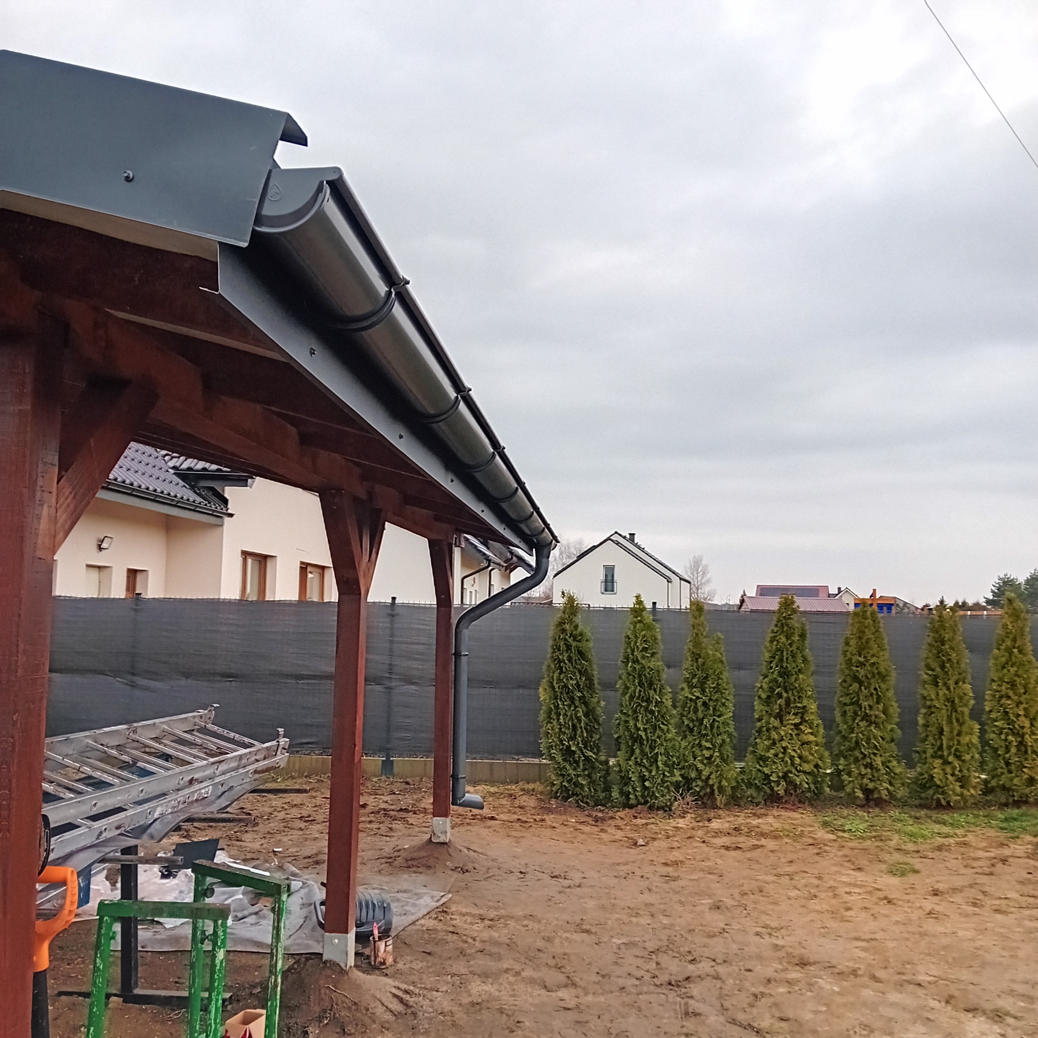 Zamontowana, grafitowa rynna na drewnianej konstrukcji w Dzierżoniowie. Widoczne narzędzia i drabina, sugerujące świeży montaż. Tło: dom i tuje.