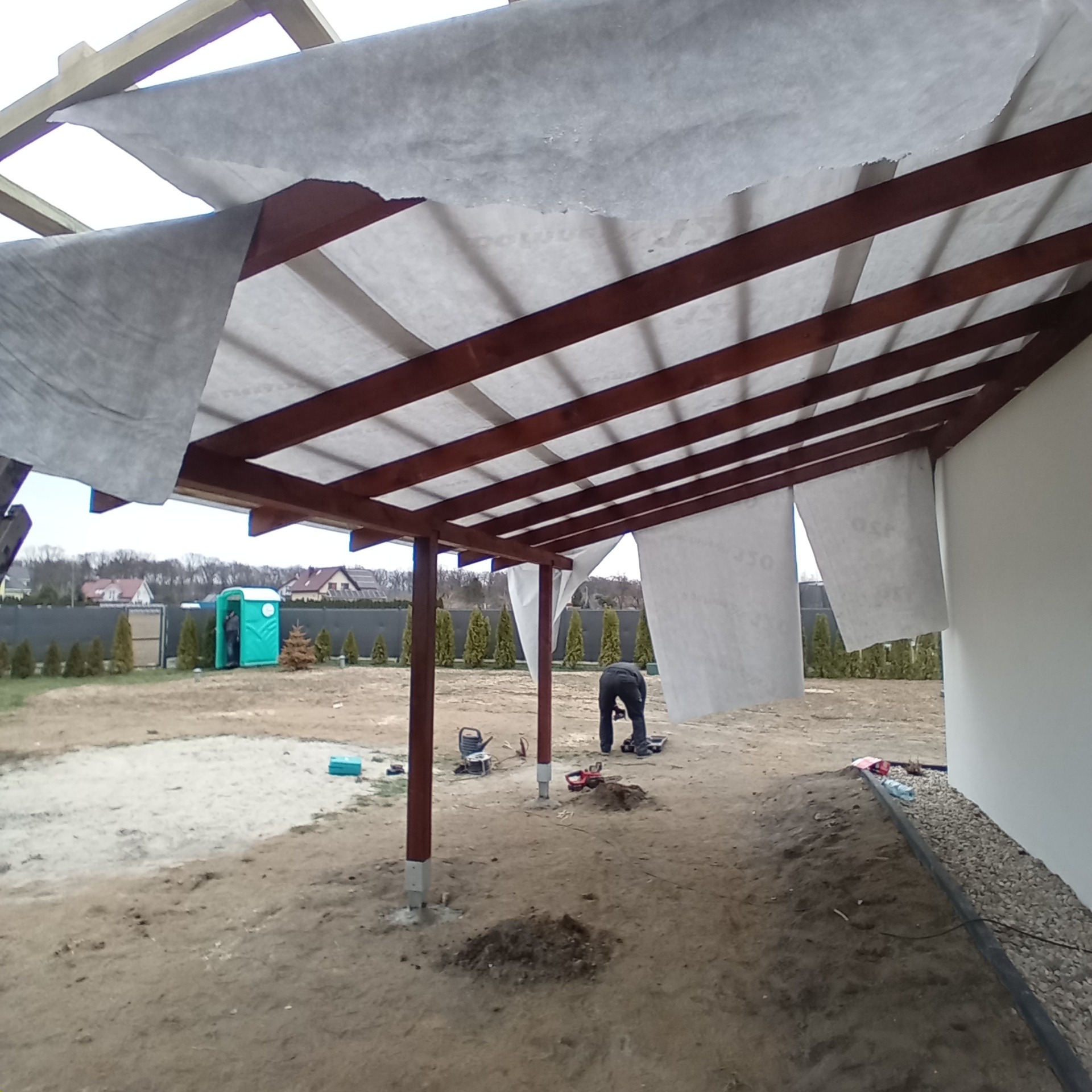 Montaż drewnianej konstrukcji dachu z membraną dachową. Widoczny pracownik w trakcie pracy, narzędzia i elementy konstrukcyjne. Ujęcie z dołu, szeroki kadr.