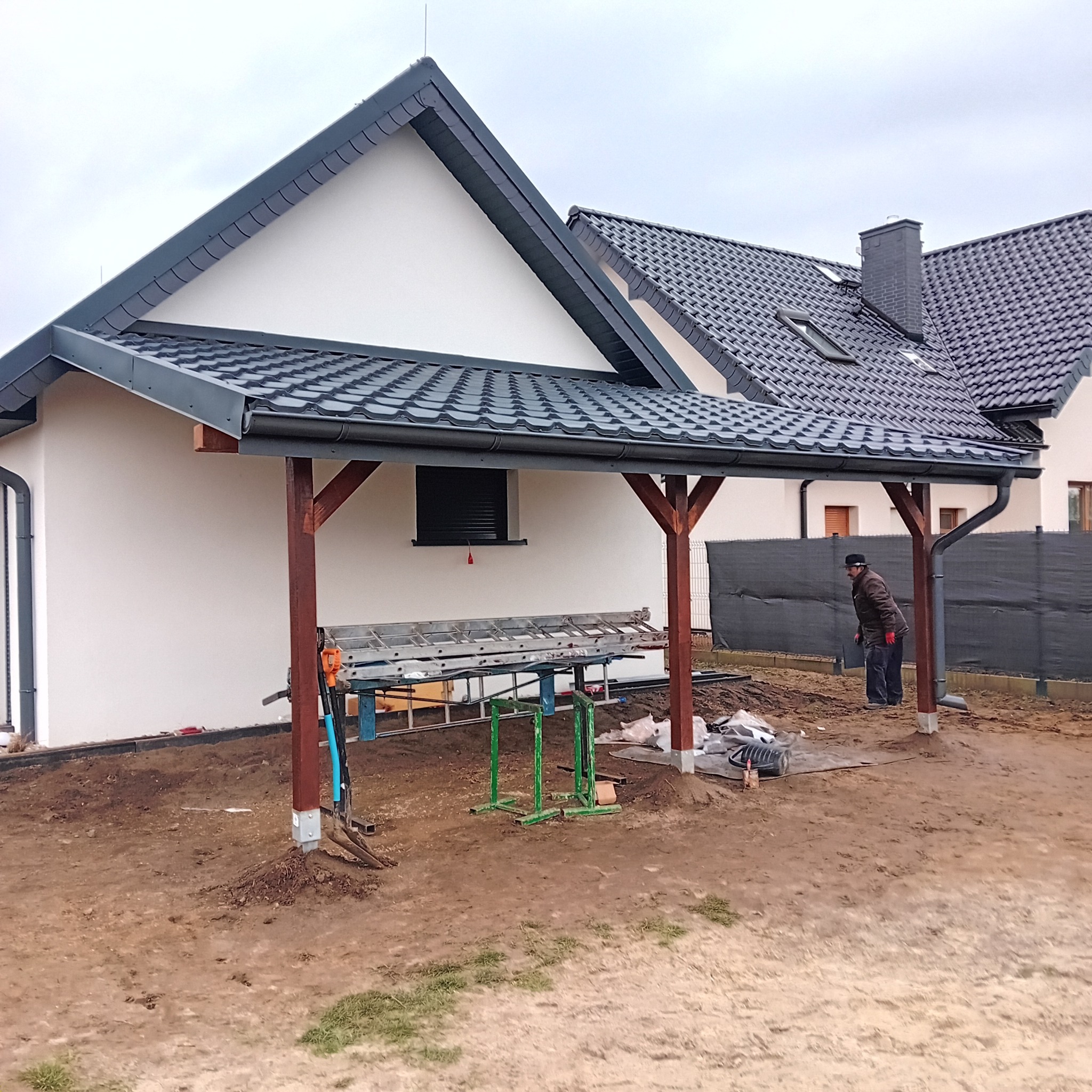 Nowoczesny dom z ciemną dachówką karpiówką i drewnianą konstrukcją zadaszenia. Widoczne rynny i obróbki blacharskie. Na pierwszym planie narzędzia i przygotowany teren.