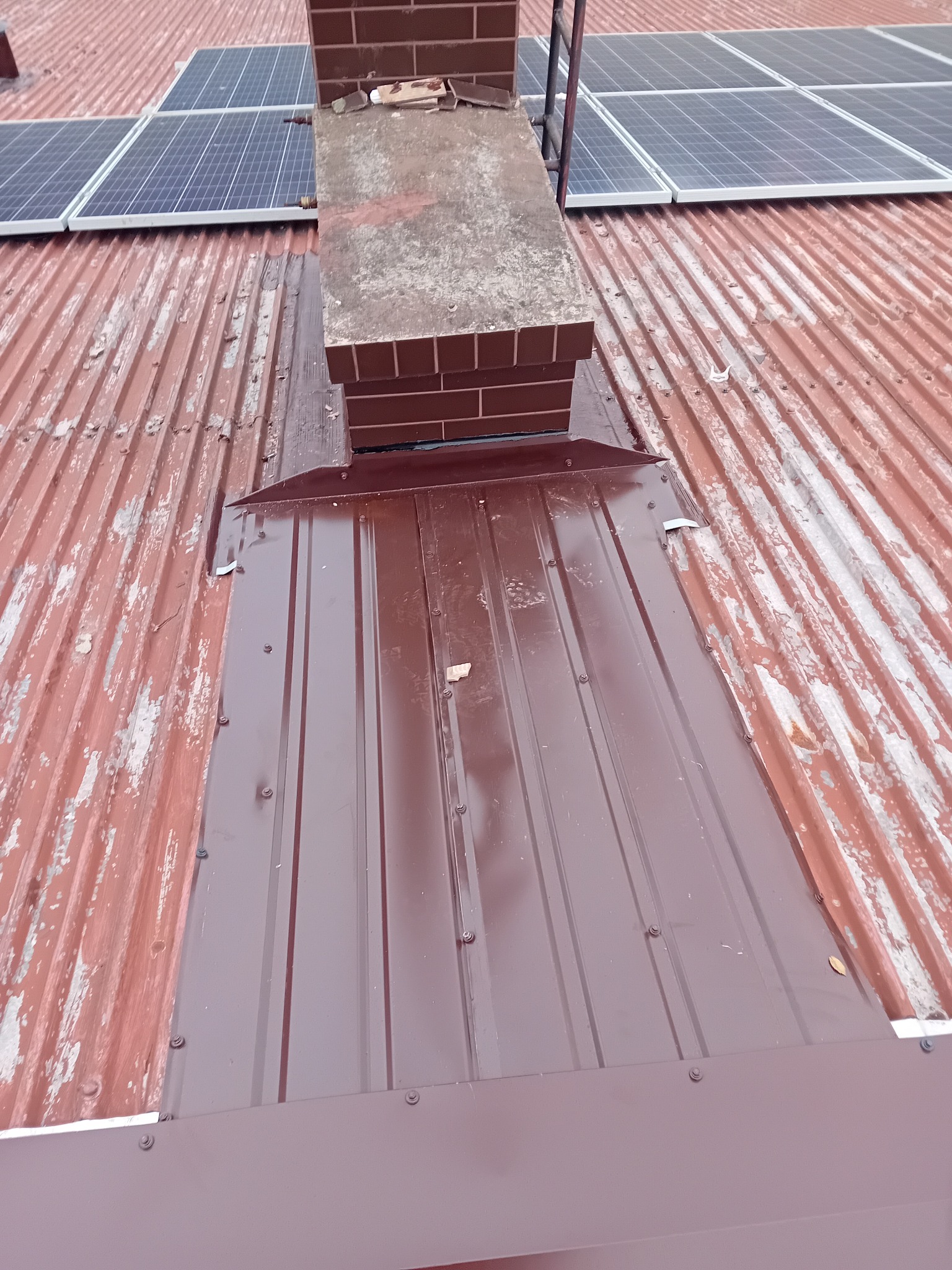 Ciemnobrązowa blacha na dachu z panelami słonecznymi i kominem. Widoczne ślady korozji na starej części dachu. Nowa blacha zamontowana przy kominie.