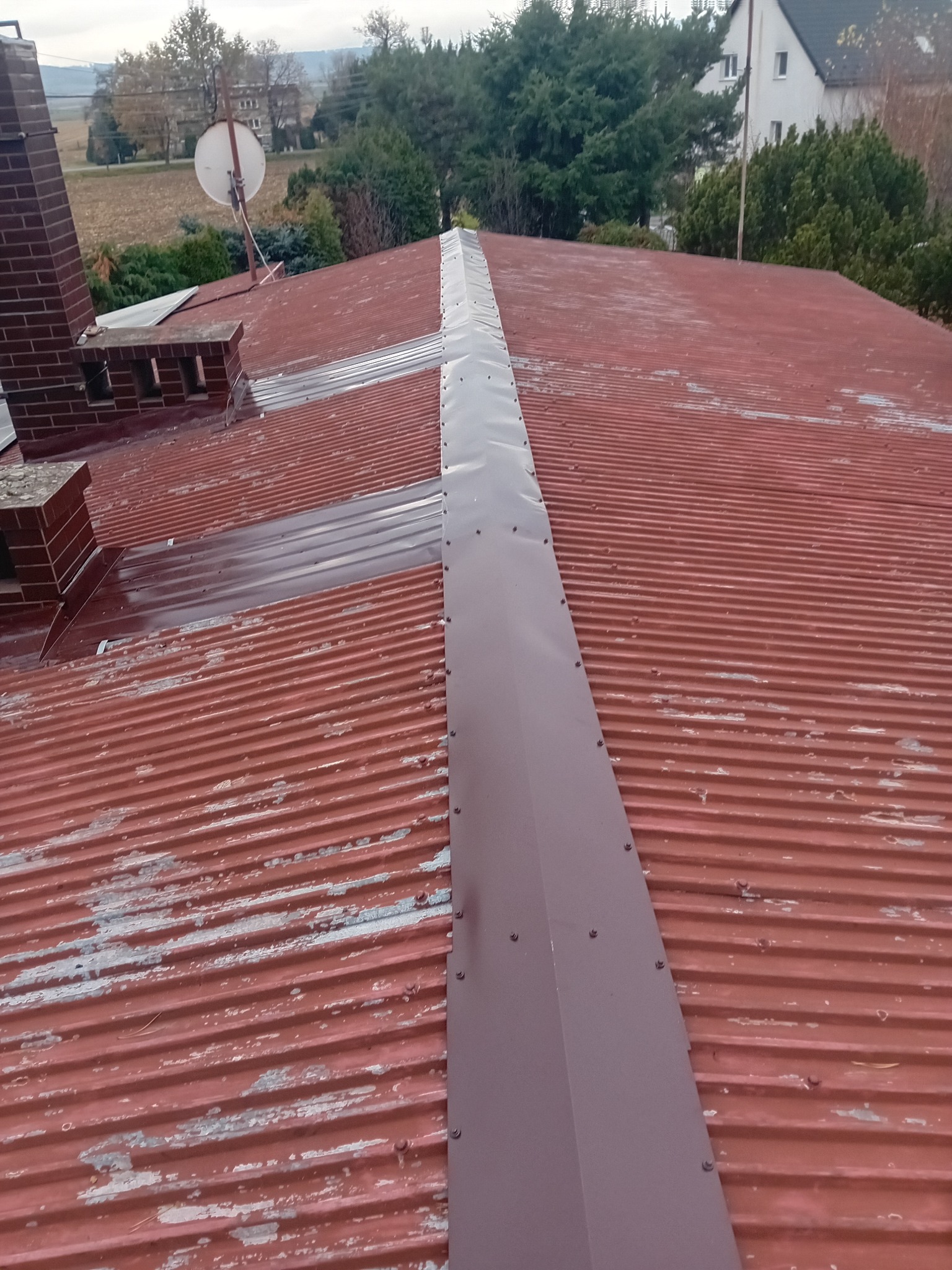 Czerwony dach z blachy trapezowej z nową, centralną obróbką blacharską w kolorze brązowym, mocowaną na nity. Widoczne ślady starej farby.