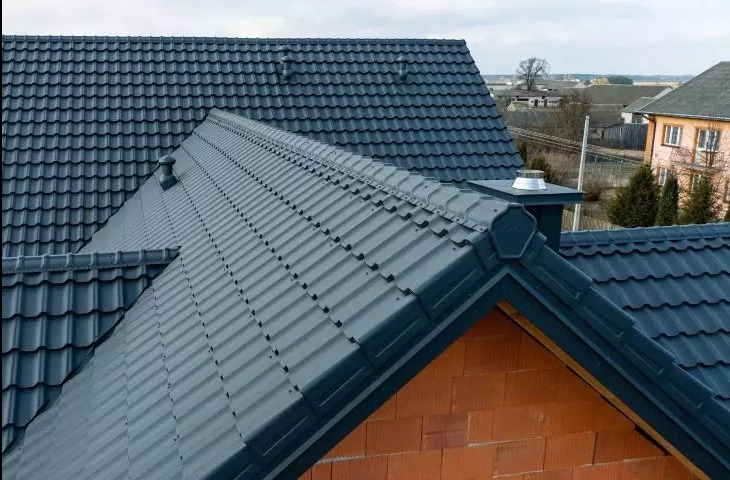 Ciemnoszary dach z blachodachówki modułowej, widok z góry na komin z wentylatorem i fragment muru z cegły ceramicznej. W tle zabudowa jednorodzinna.