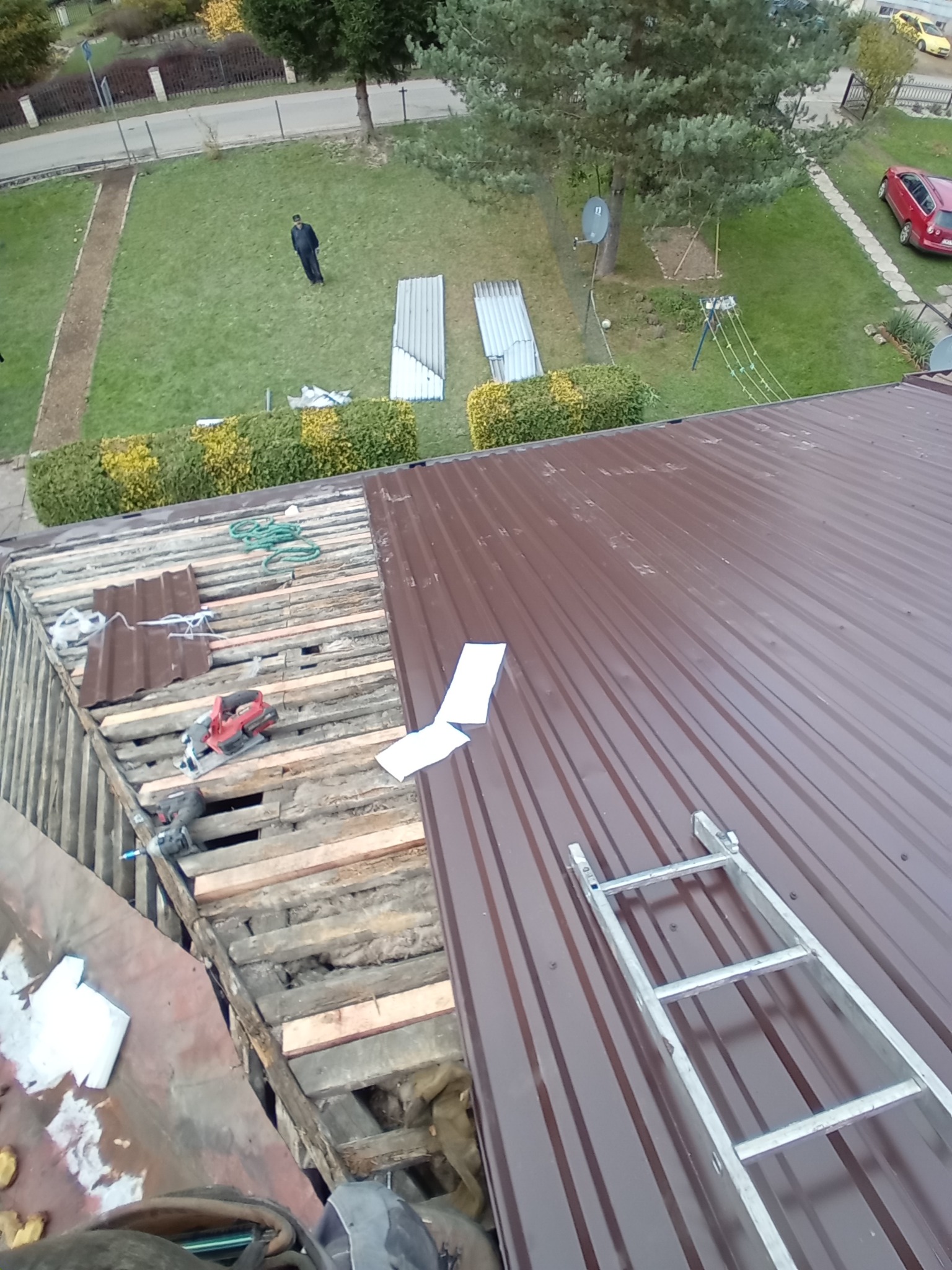 Częściowo zdjęte brązowe pokrycie dachu z widoczną konstrukcją drewnianą i ociepleniem. Na dachu leżą narzędzia i drabina. W tle trawnik z osobą i blachą.