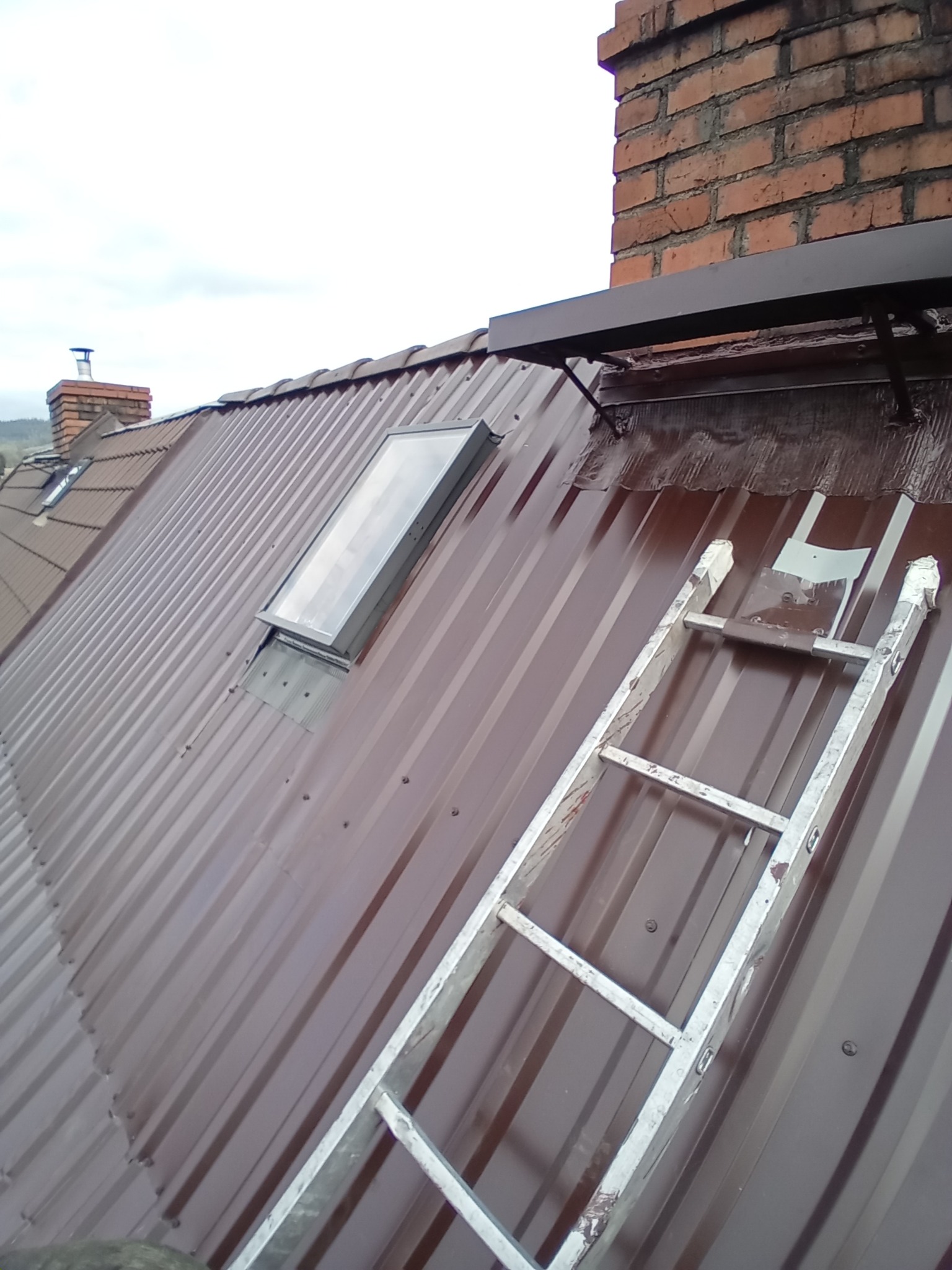 Brązowy dach z blachy trapezowej z oknem dachowym i kominem z cegły, częściowo zasłonięty drabiną. Widok z góry na nierówny teren w tle.
