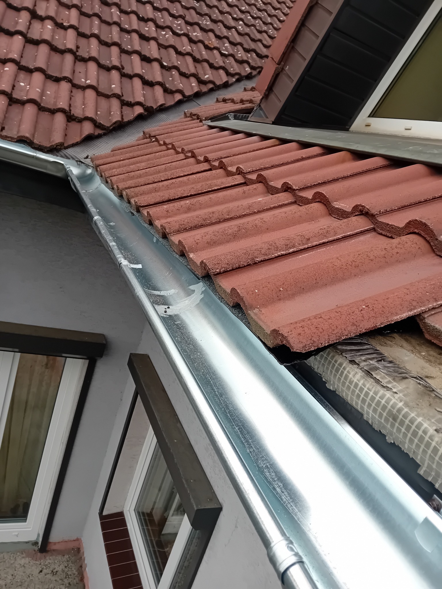 Błyszcząca, nowa rynna zamontowana na tle czerwonej dachówki i fragmentu okna. Widoczne mocowania rynny. Ujęcie z dołu, podkreślające detale instalacji.
