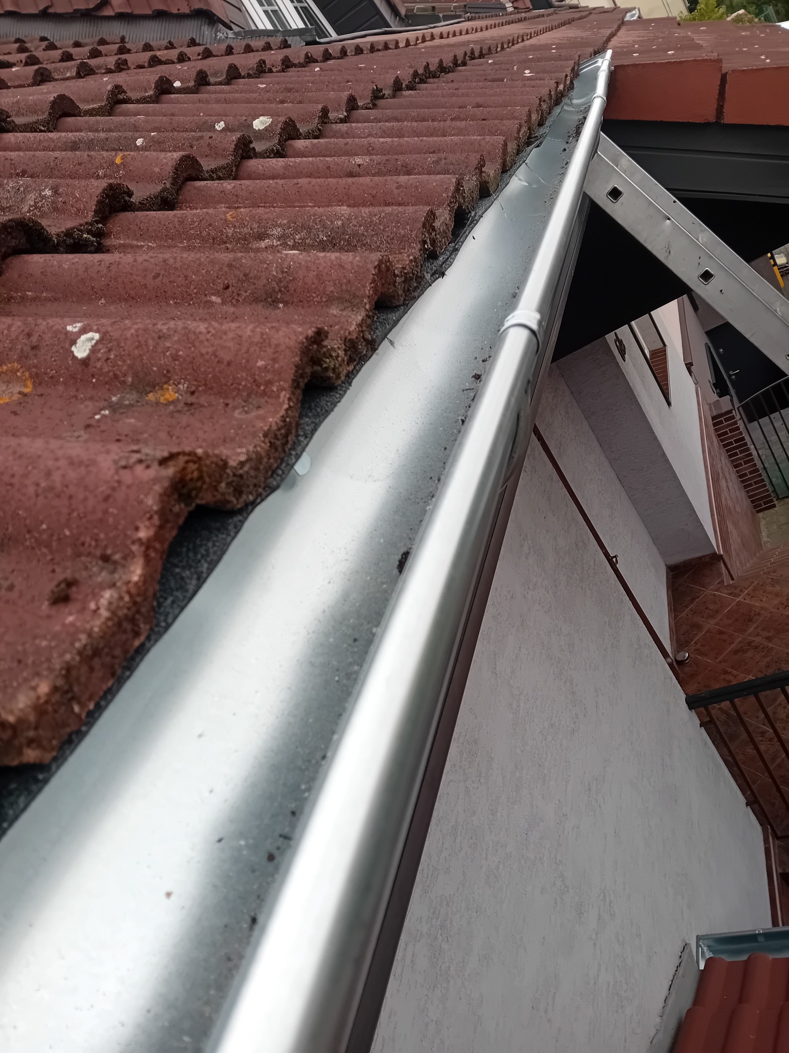 Nowa, błyszcząca rynna zamontowana na dachu z czerwonej dachówki. Widoczny fragment drabiny oparty o ścianę budynku. Ujęcie z dołu, podkreślające detale.