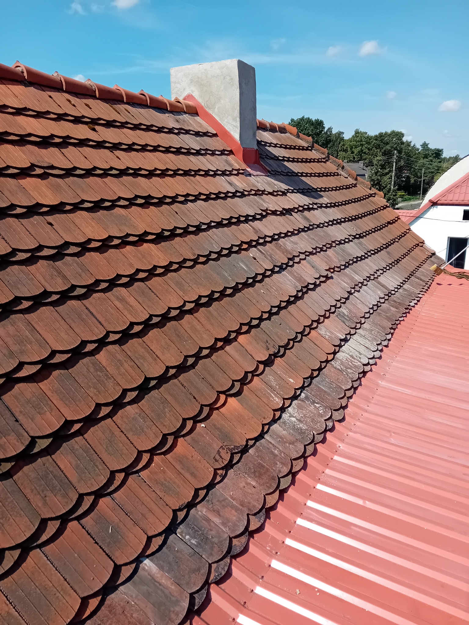 Czerwony dach z dachówki karpiówki i blachy trapezowej, widok z góry, komin w tle, kontrast czystej i zabrudzonej powierzchni, błękitne niebo.