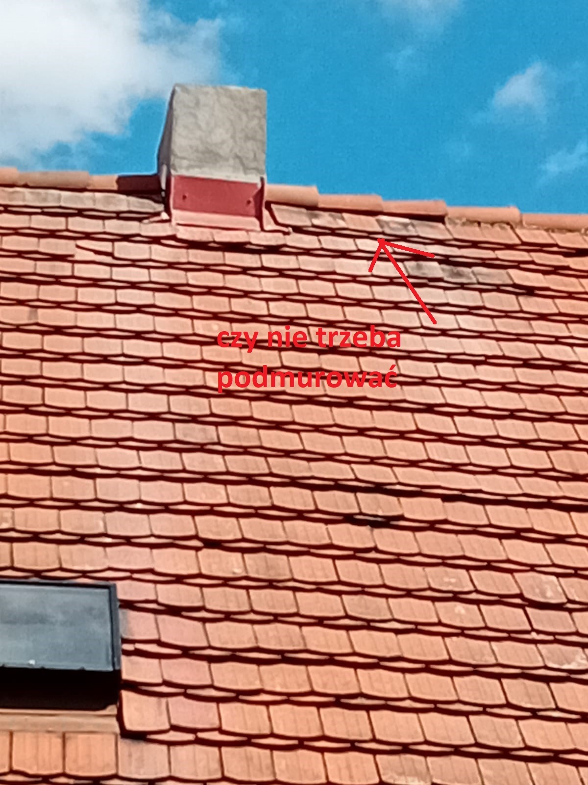 Ceglasty dach z kominem i fragmentem okna dachowego, widoczne zabrudzenia na dachówkach. Na zdjęciu naniesiono tekst i strzałkę.