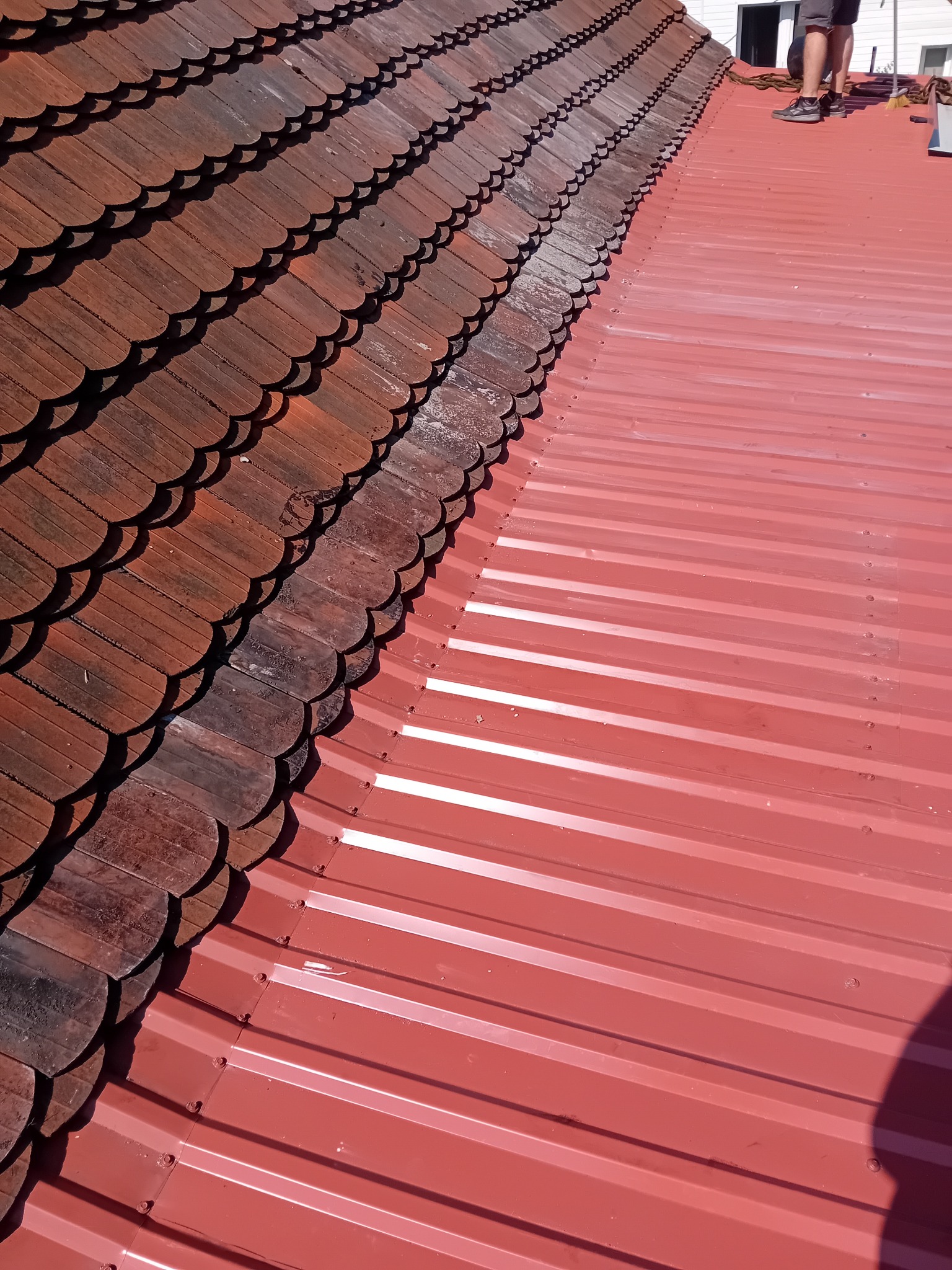 Częściowo odnowiony dach: stara, łuskowata dachówka obok nowej, czerwonej blachy trapezowej. Widoczne mocowania blachy. Praca w toku, fragment osoby w kadrze.