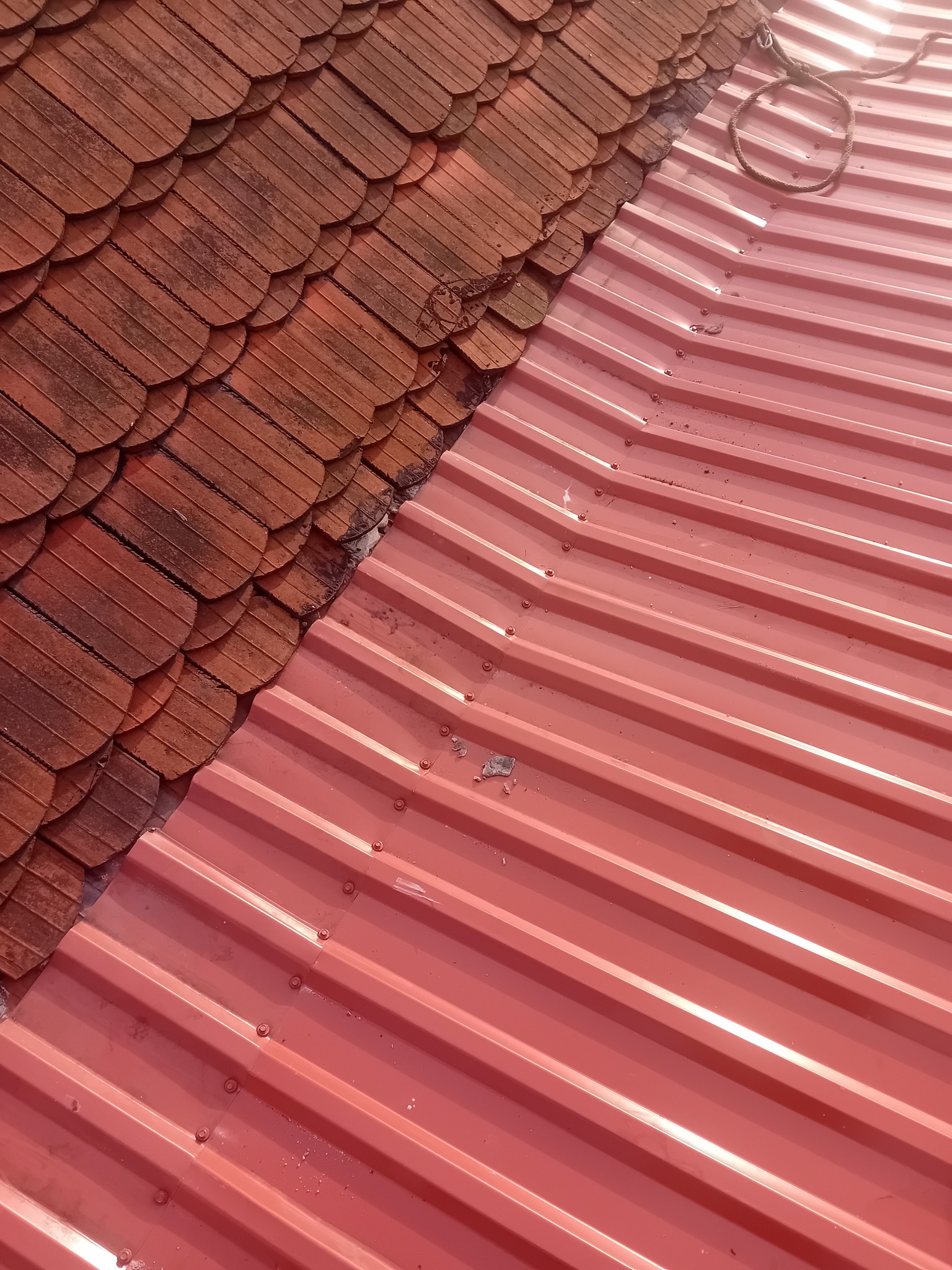 Czerwona blacha trapezowa na dachu, kontrastująca z fragmentem starej, brązowej dachówki karpiówki. Widoczne mocowania blachy. Ujęcie z góry, ukośne.