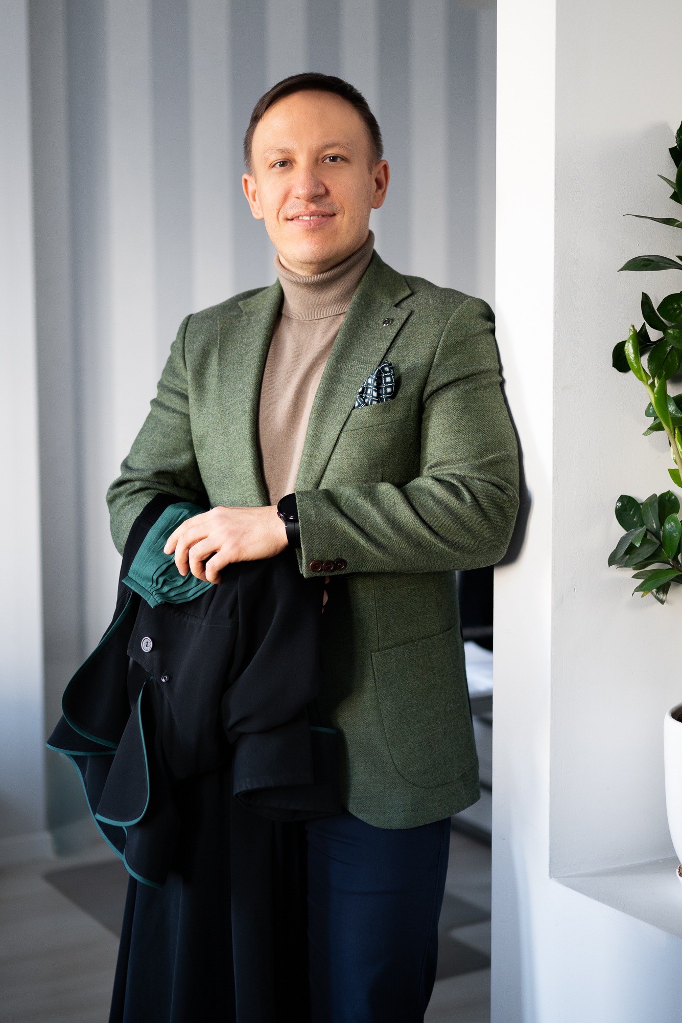 Uśmiechnięty mężczyzna w zielonej marynarce trzyma togę adwokacką na tle białych ścian i rośliny doniczkowej. Elegancki strój, profesjonalny wizerunek.