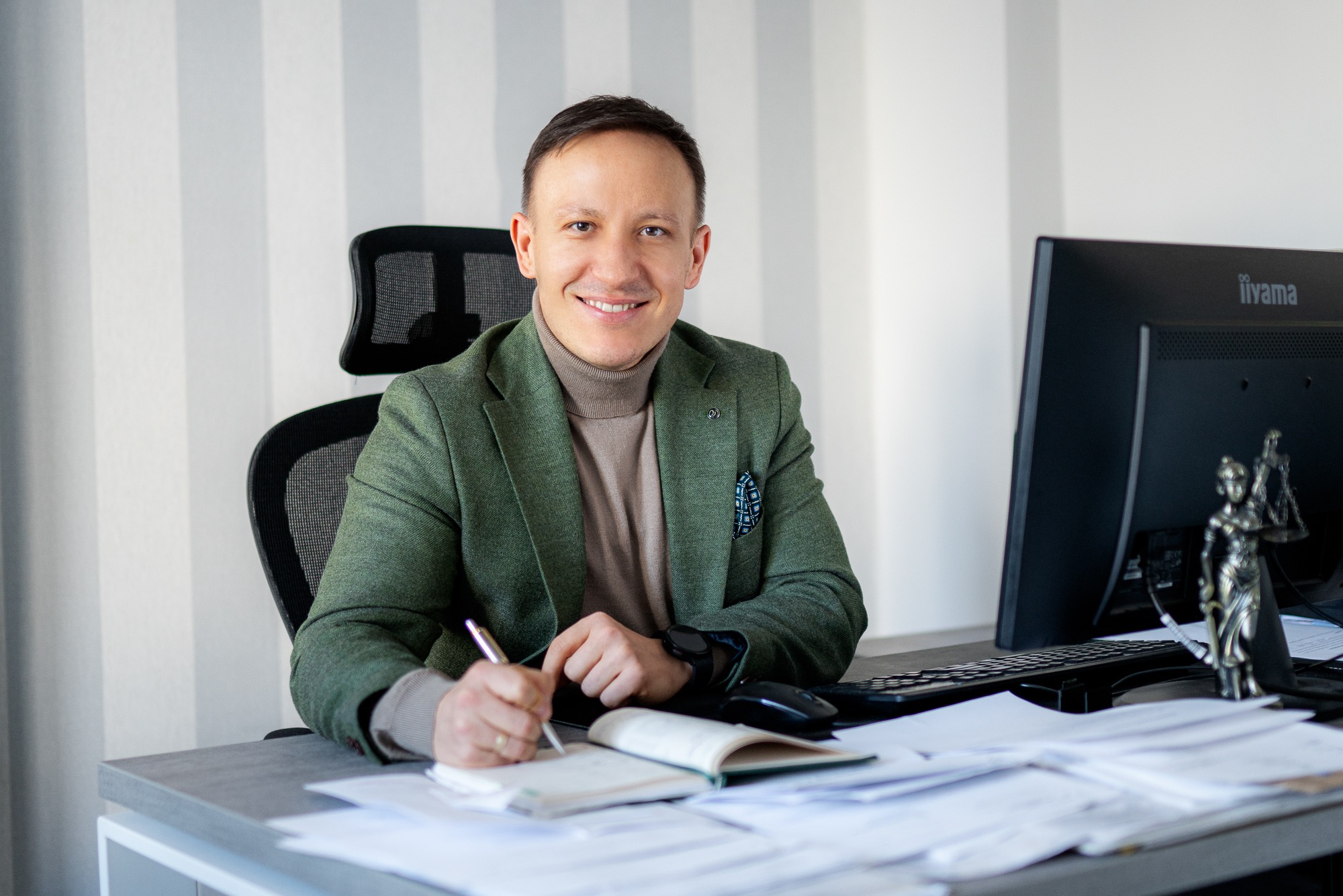 Uśmiechnięty mężczyzna w zielonej marynarce siedzi za biurkiem, pisząc w notatniku. Na biurku komputer, papiery i figurka Temidy. Tło stanowią pionowe pasy na ścianie.