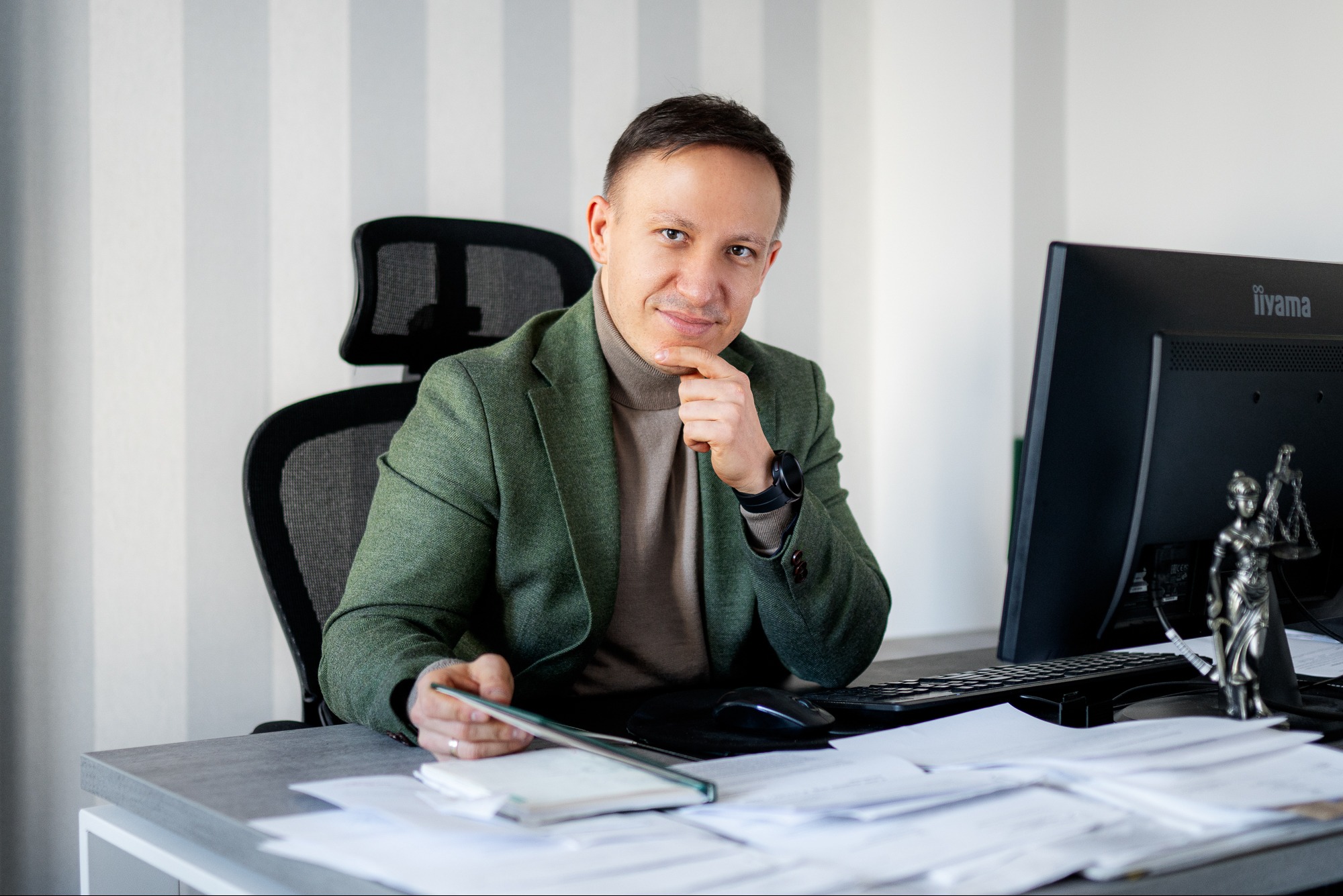 Portret mężczyzny w zielonej marynarce siedzącego za biurkiem z komputerem i figurką Temidy. Uśmiechnięty, zamyślony, w tle ściana w pionowe pasy.