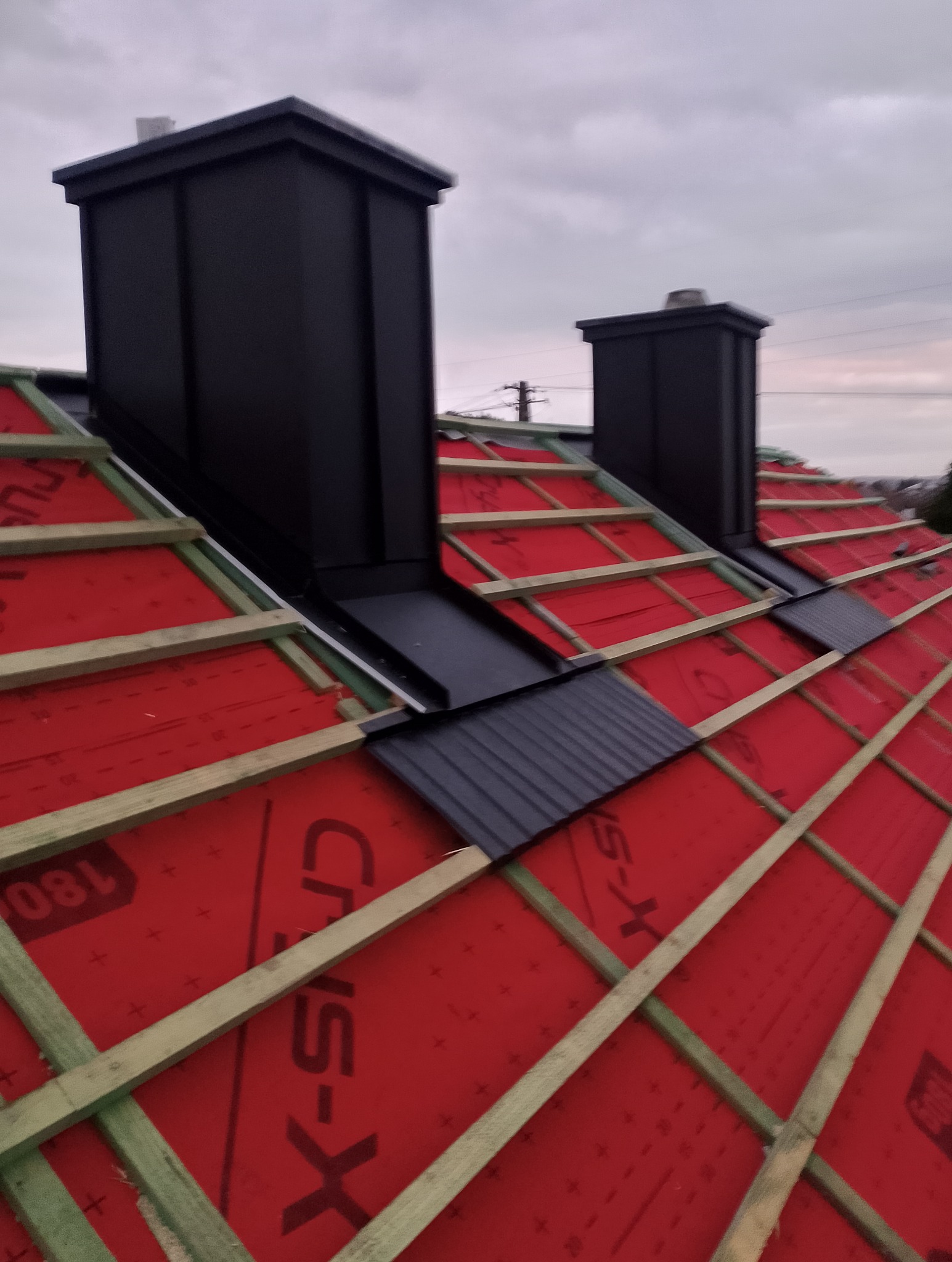 Częściowo pokryty dach z czarnymi kominami i czerwoną membraną dachową. Widoczne drewniane łaty. Ujęcie z góry, pochmurne niebo w tle.