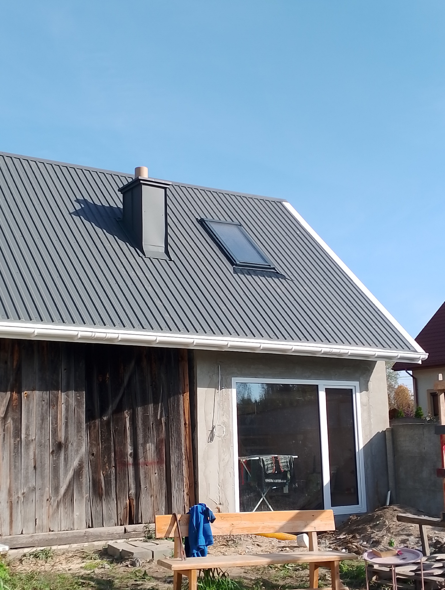 Szary dach z blachy trapezowej z oknem dachowym i kominem. Widoczne białe rynny i drewniana brama garażowa. W tle błękitne niebo.