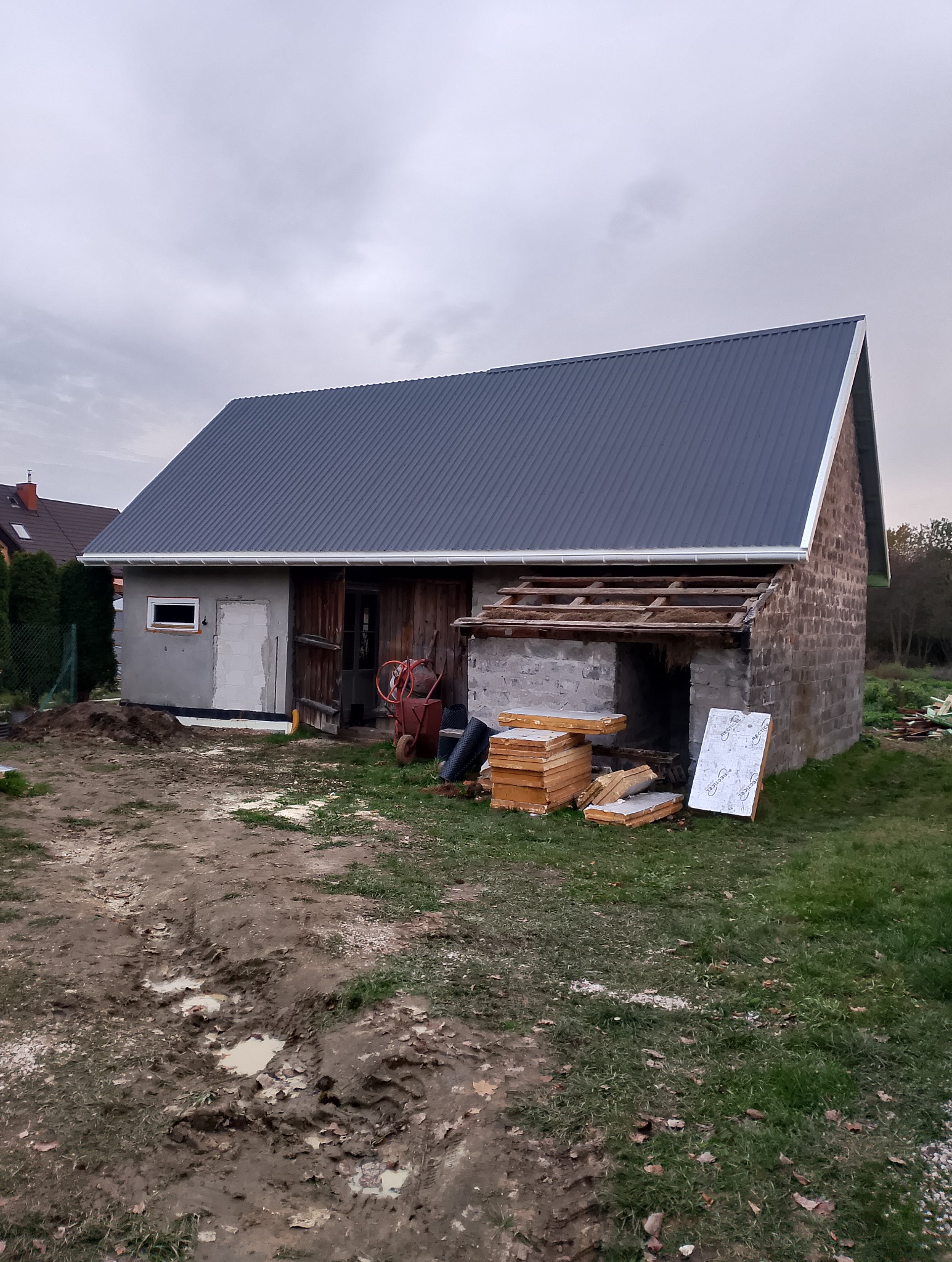 Budynek gospodarczy z częściowo wymienionym dachem z blachy trapezowej. Widoczne materiały budowlane i rusztowanie. Część dachu jeszcze bez pokrycia.