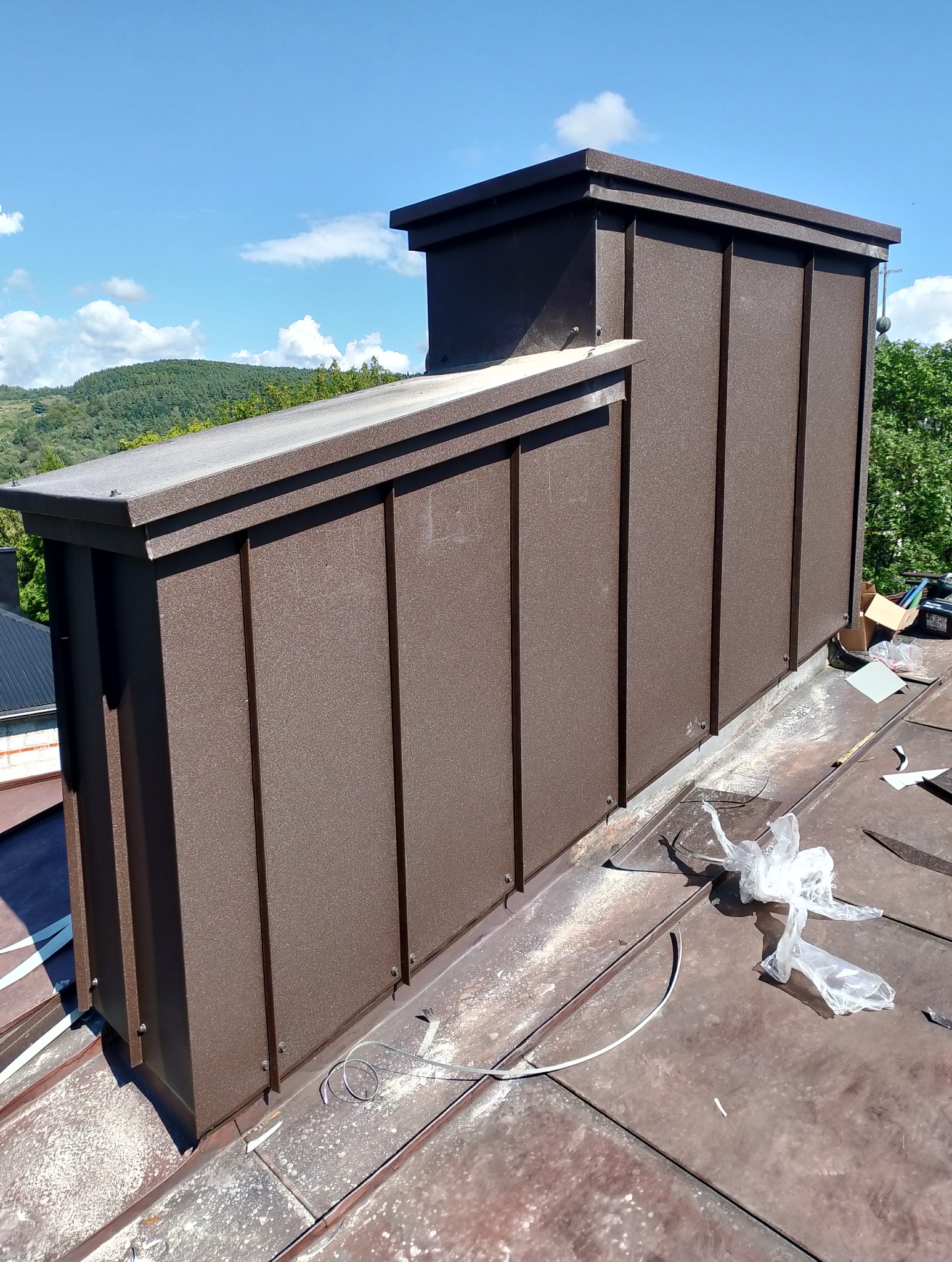 Nowoczesna obudowa komina z brązowej blachy na dachu, widok z góry, w tle zalesione wzgórza i błękitne niebo. Detal obróbki blacharskiej.