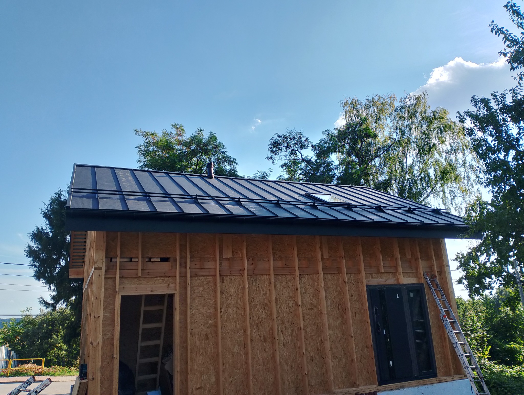 Nowoczesny, ciemny dach z blachy na budynku w trakcie budowy, widoczne deski i okno. Na tle błękitne niebo i drzewa. Dach z oknem dachowym.