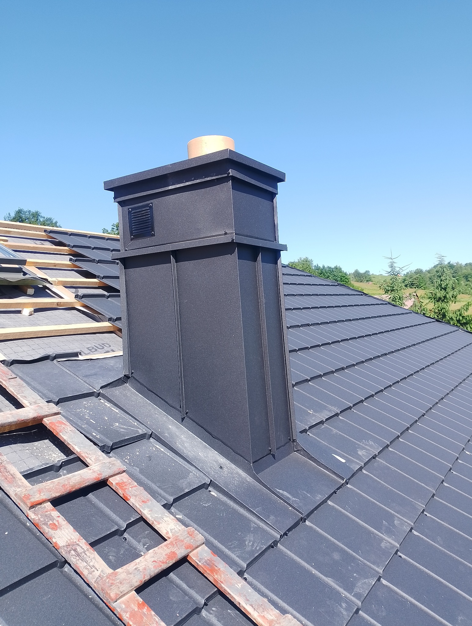Nowoczesny, grafitowy komin z blachy na dachu z dachówki modułowej, fragment rusztowania dekarskiego, błękitne niebo w tle. Detal obróbki blacharskiej.