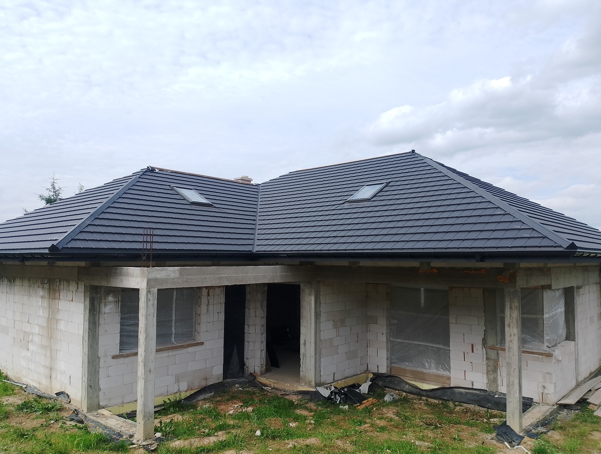 Dom w trakcie budowy z nowym, grafitowym dachem z dachówki ceramicznej i oknami połaciowymi. Widoczne rynny i obróbki blacharskie.