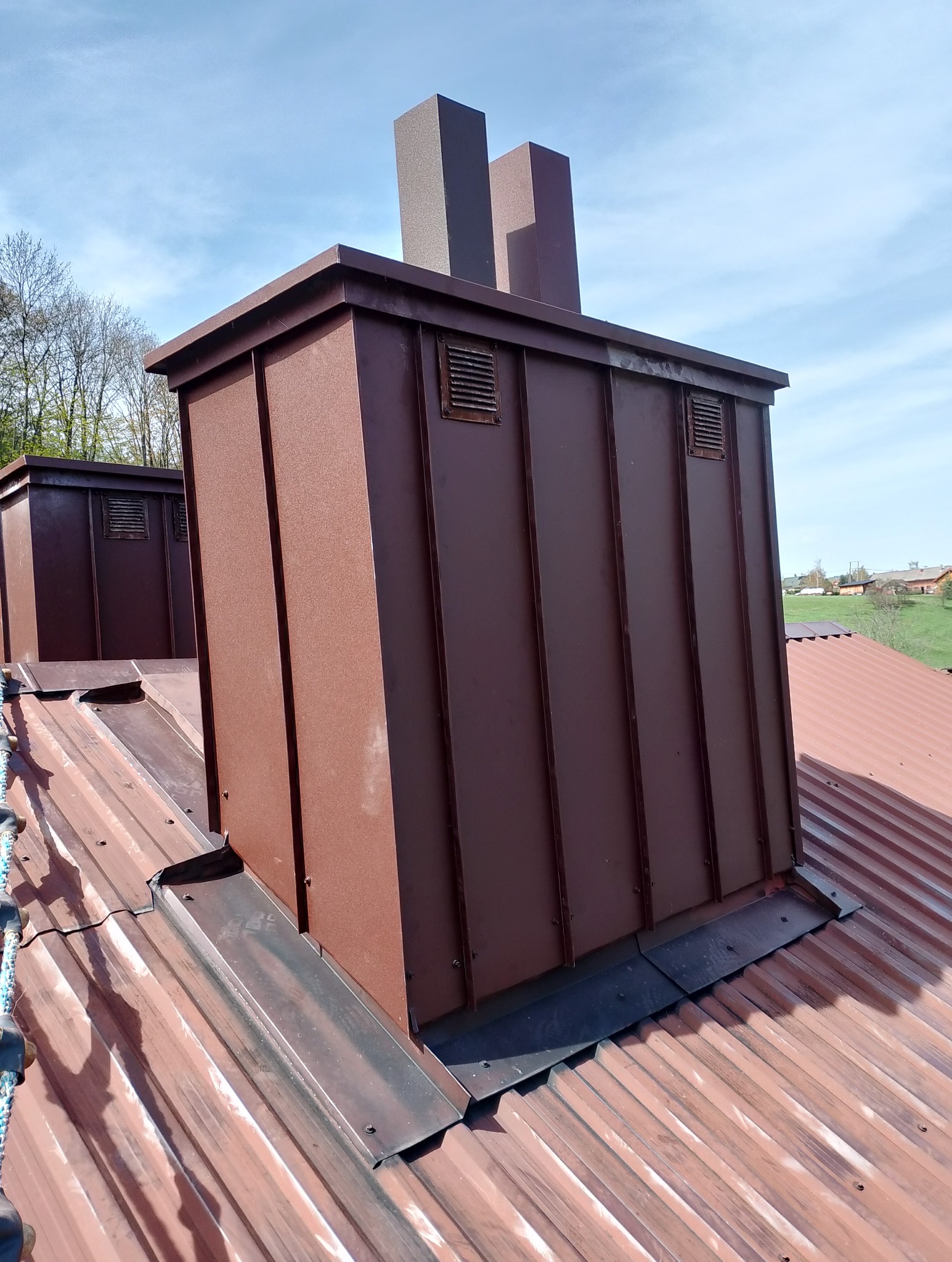 Brązowe obróbki blacharskie komina wentylacyjnego na dachu z blachy trapezowej w słoneczny dzień. Widoczne detale łączeń i wentylacji.