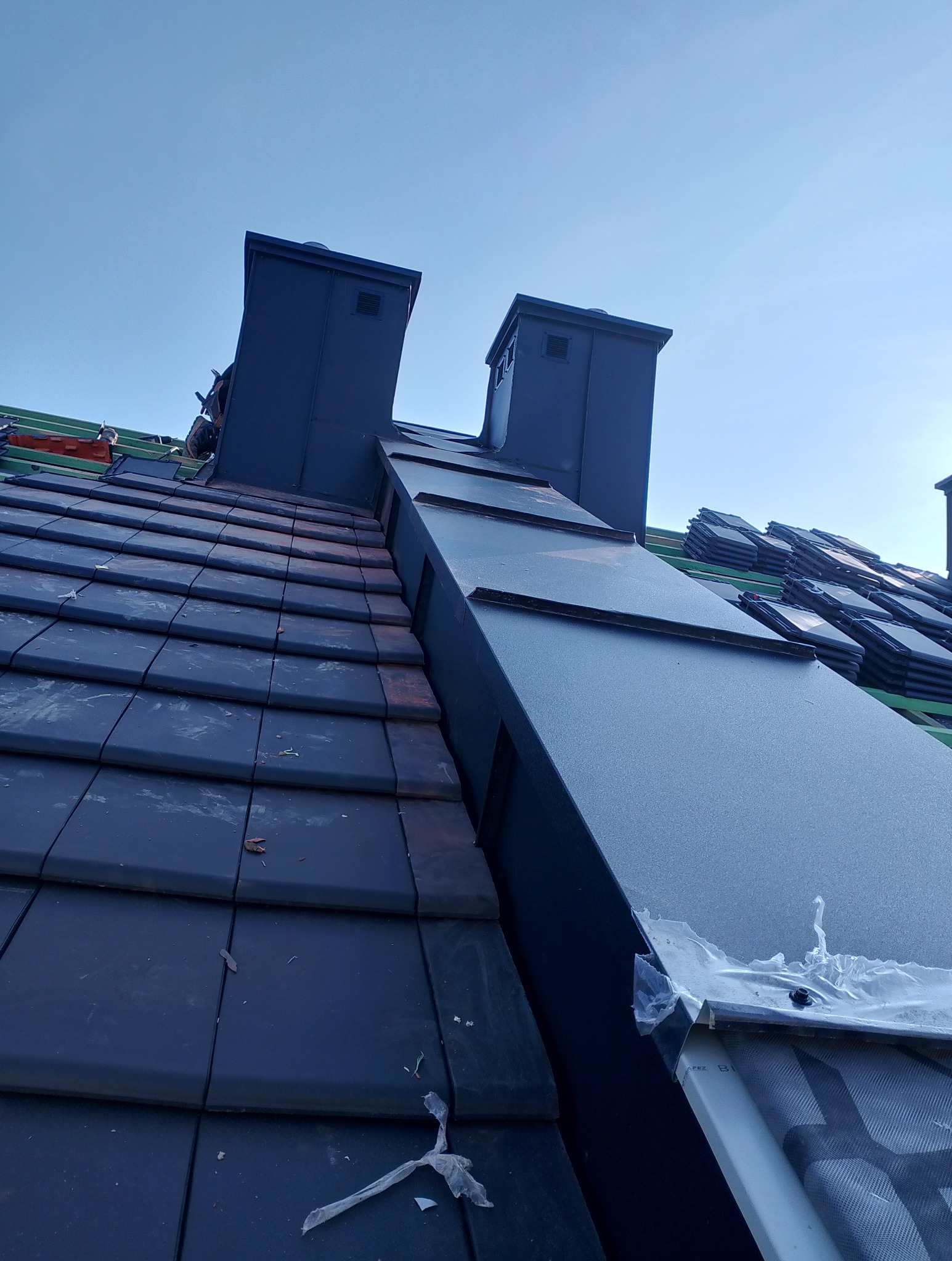 Częściowo pokryty dach z ciemnej dachówki ceramicznej i blachy, widoczne kominy. Na dachu leżą stosy dachówek. Ujęcie z dołu, niebo w tle.