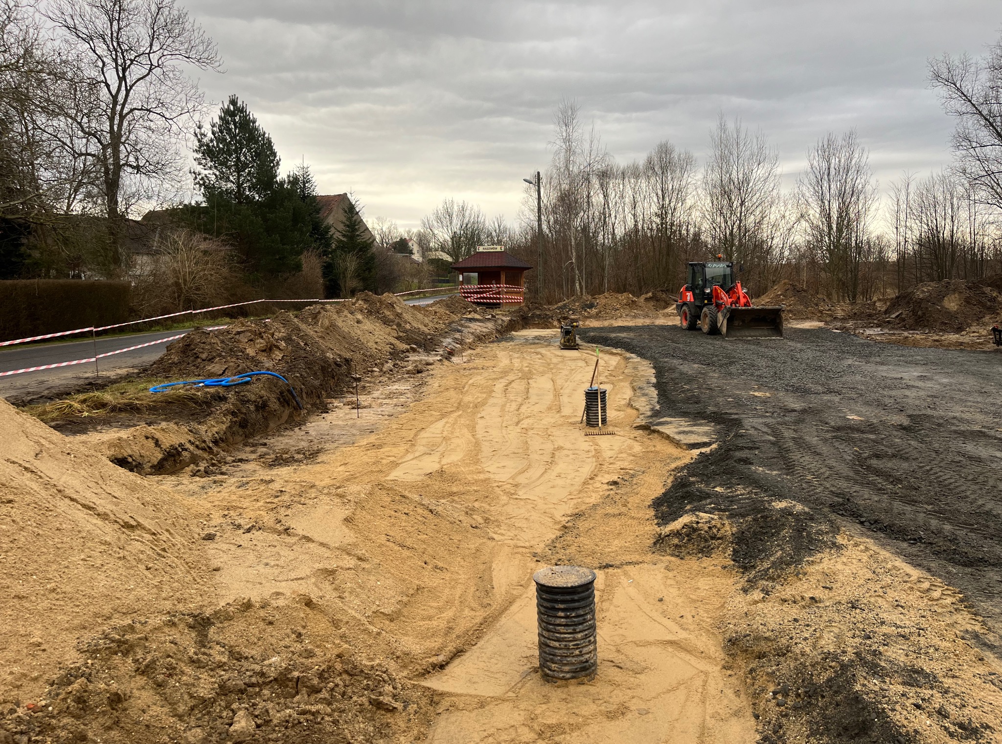 Wykonane wykopy pod instalacje na posesji, widoczny sprzęt budowlany i studzienki kanalizacyjne. Prace ziemne w toku, teren zabezpieczony taśmą ostrzegawczą.