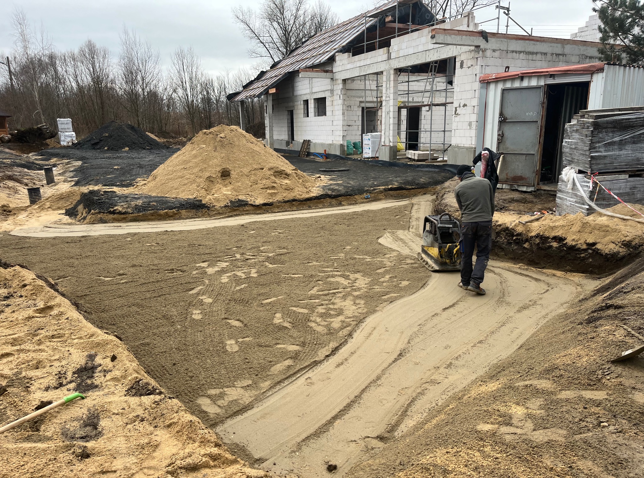 Prace ziemne: ubijanie piasku zagęszczarką na budowie domu jednorodzinnego. W tle budynek w stanie surowym otwartym i hałdy piasku. Widoczne narzędzia i pracownik.