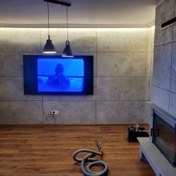 Fidar design - Nowoczesne wnętrze z panelami ściennymi imitującymi beton, drewnianą podłogą i minimalistycznym kominkiem. Dwie lampy wiszące nad telewizorem. Wąż od odkurzacza na podłodze.