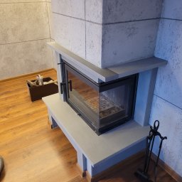 Fidar design - Nowoczesny kominek z szarą obudową i czarnym wkładem, drewniana podłoga, ściana o fakturze betonu. Widoczne akcesoria kominkowe i odkurzacz.