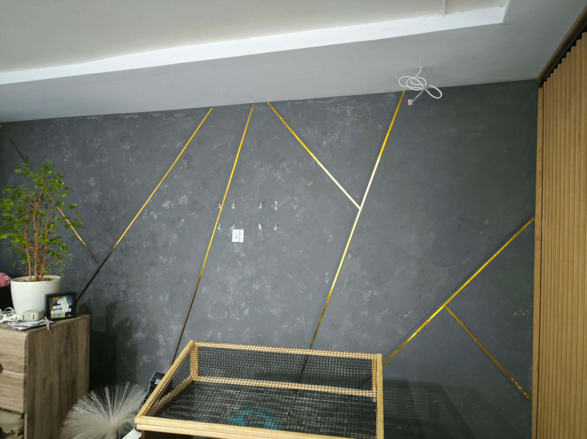 Dekoracyjna ściana w szarościach z geometrycznym wzorem ze złotych pasków. Widoczny fragment drewnianej boazerii i rośliny doniczkowej. Nowoczesny design wnętrza.