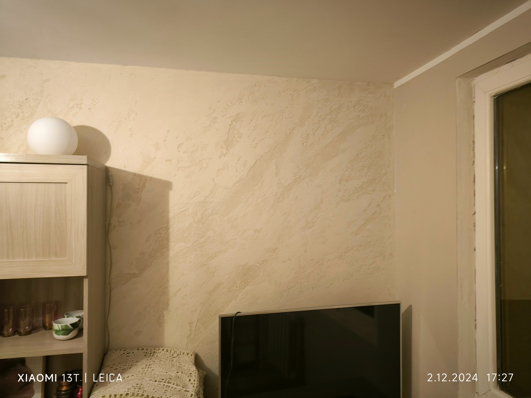 Ściana z dekoracyjną fakturą w odcieniach beżu, oświetlona lampą. Widoczny fragment szafki i telewizora. Efektowna aranżacja wnętrza.