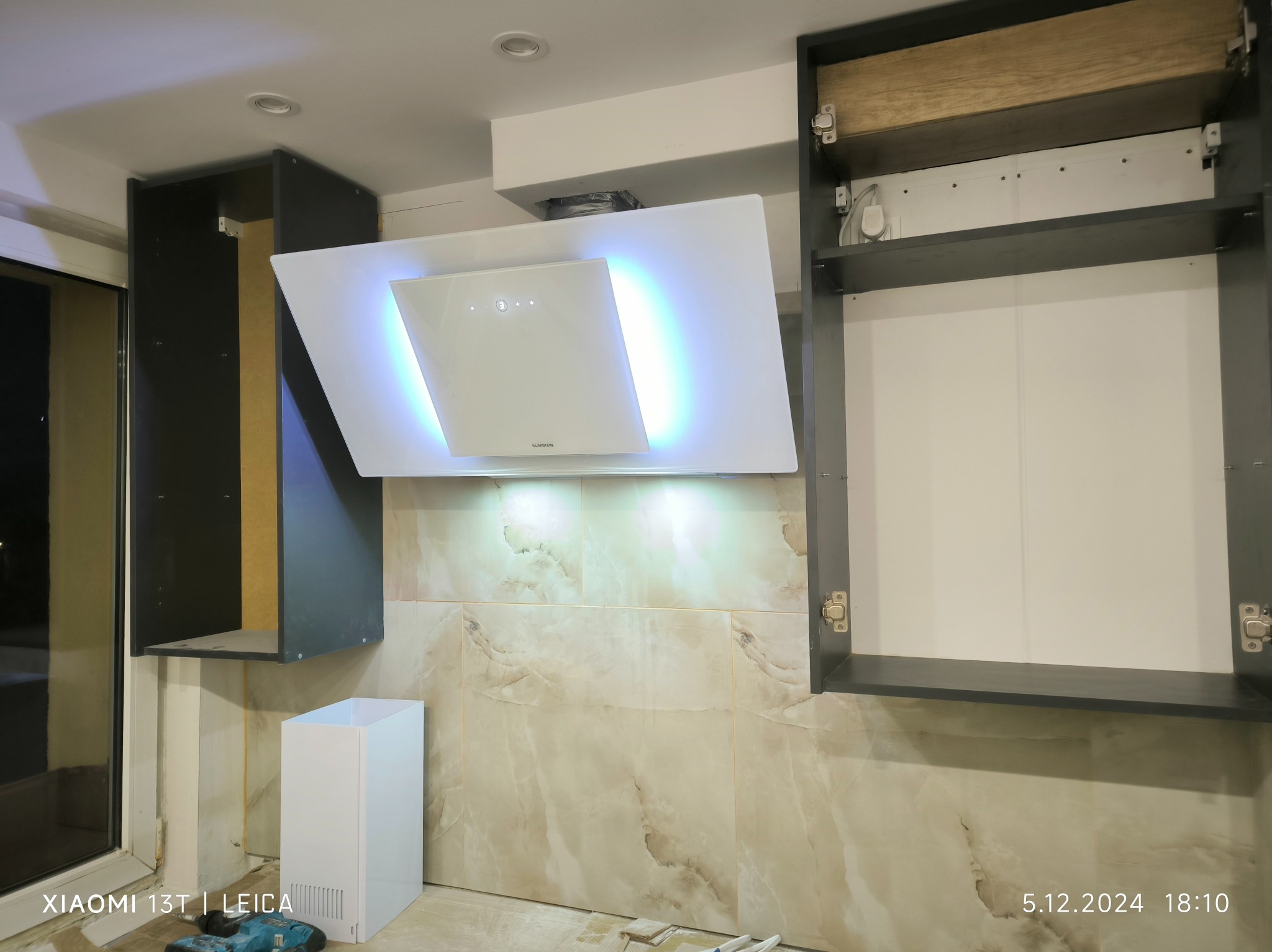 Montaż okapu kuchennego Klarstein z niebieskim podświetleniem LED, widoczne szafki kuchenne w trakcie instalacji, płytki imitujące marmur na ścianie, narzędzia na podłodze.