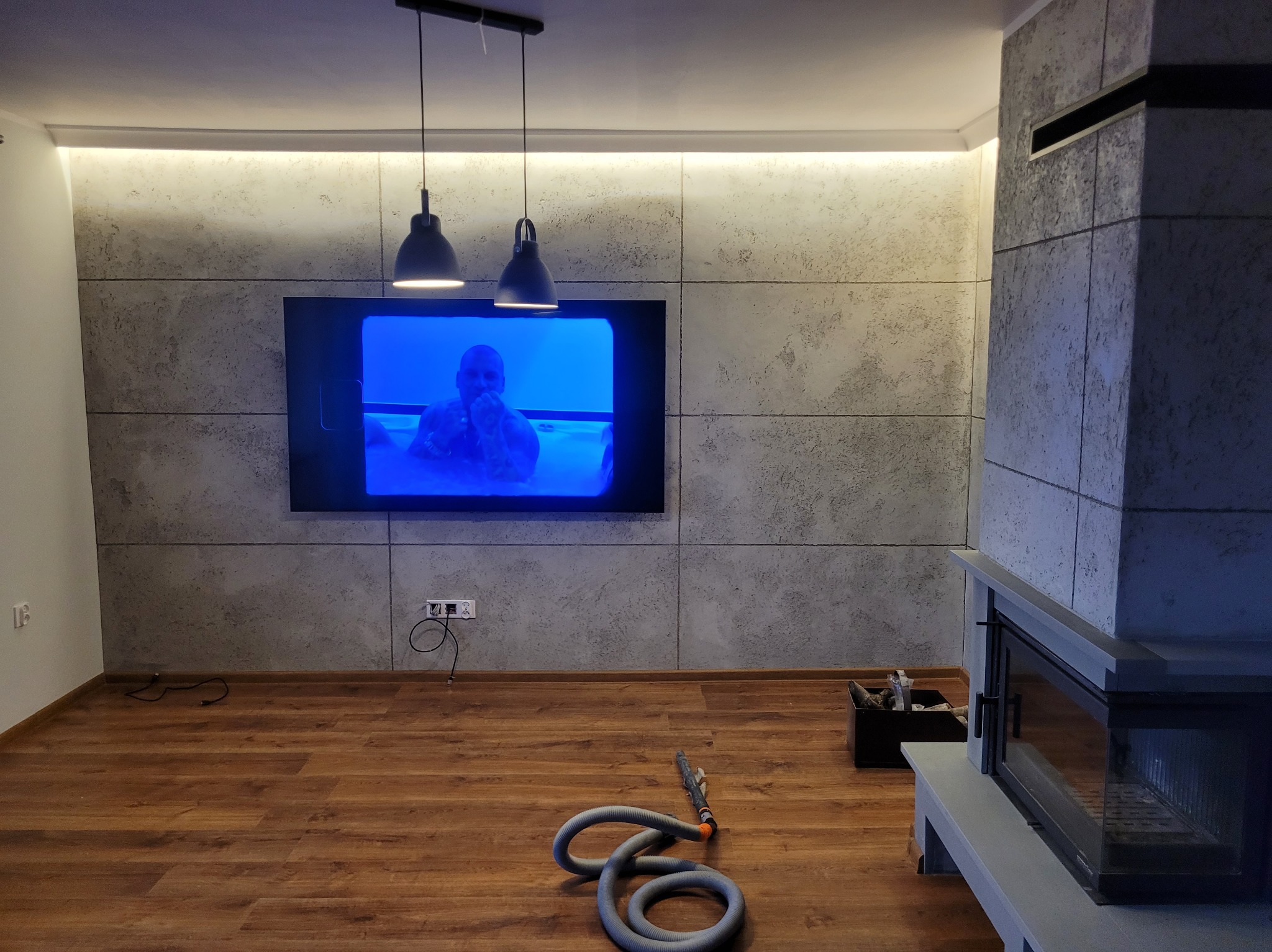 Nowoczesne wnętrze z panelami ściennymi imitującymi beton, drewnianą podłogą i minimalistycznym kominkiem. Dwie lampy wiszące nad telewizorem. Wąż od odkurzacza na podłodze.