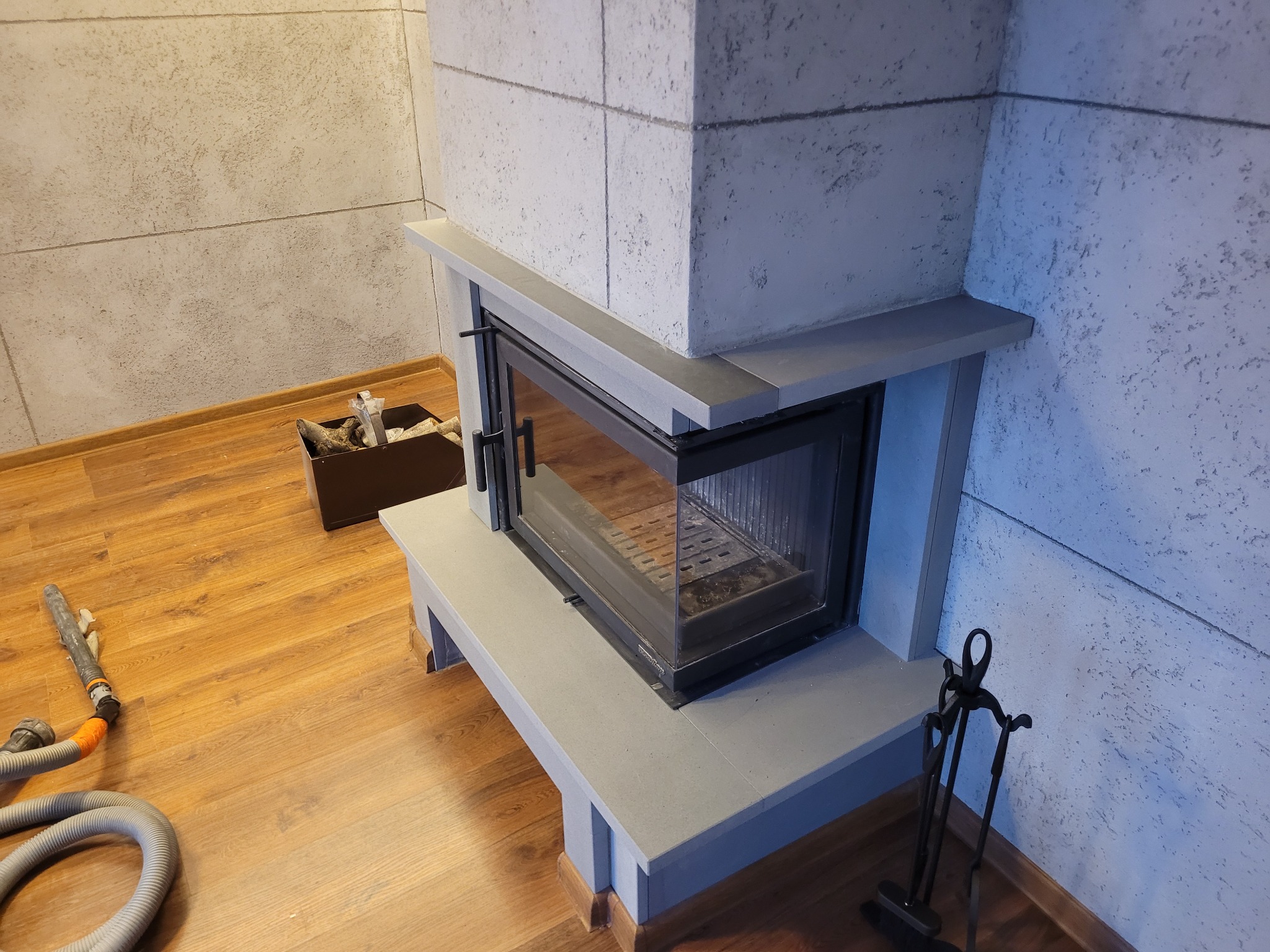 Nowoczesny kominek z szarą obudową i czarnym wkładem, drewniana podłoga, ściana o fakturze betonu. Widoczne akcesoria kominkowe i odkurzacz.