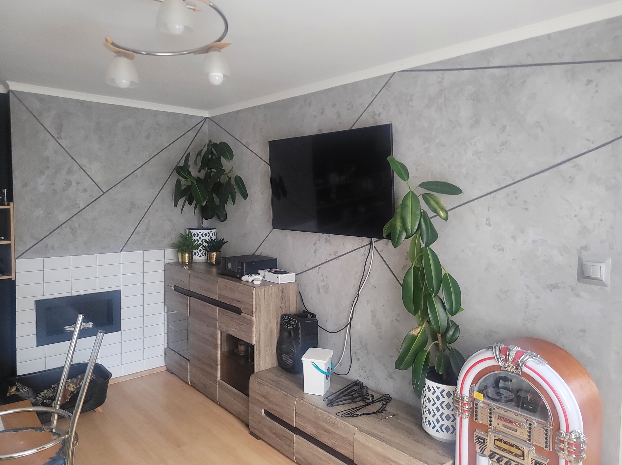 Szara ściana z geometrycznym wzorem w salonie, drewniana komoda pod telewizorem, rośliny doniczkowe i dekoracyjny jukebox. Widoczny fragment kominka z białymi płytkami.