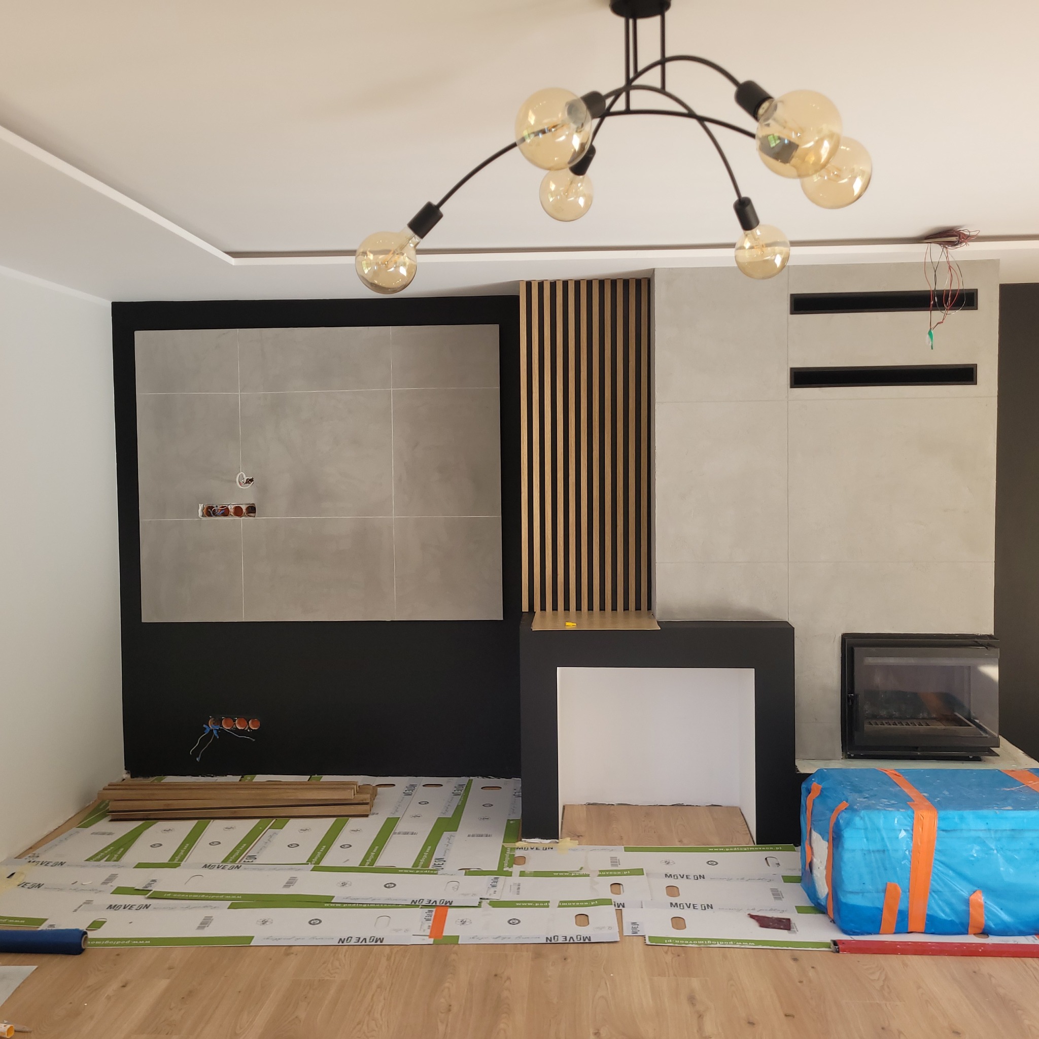 Wnętrze w trakcie prac: ułożona podkładka pod panele, widoczne panele, fragment ściany z płytkami i drewnianymi lamelami, nowoczesna lampa sufitowa. Etap prac wykończeniowych.