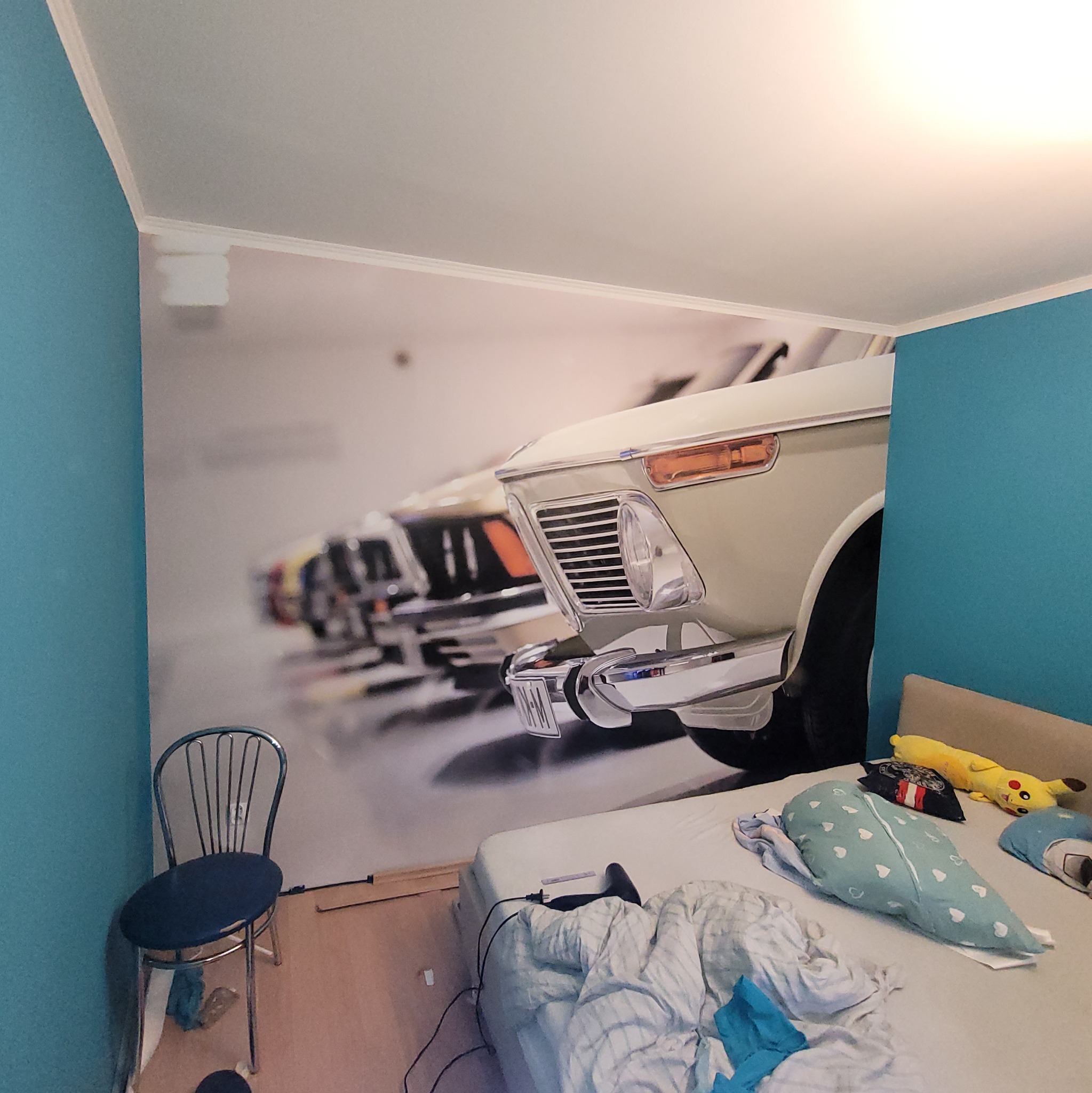 Pokój z fototapetą przedstawiającą klasyczne samochody. Niebieskie ściany, łóżko z pościelą i krzesło. Fototapeta dodaje wnętrzu charakteru.