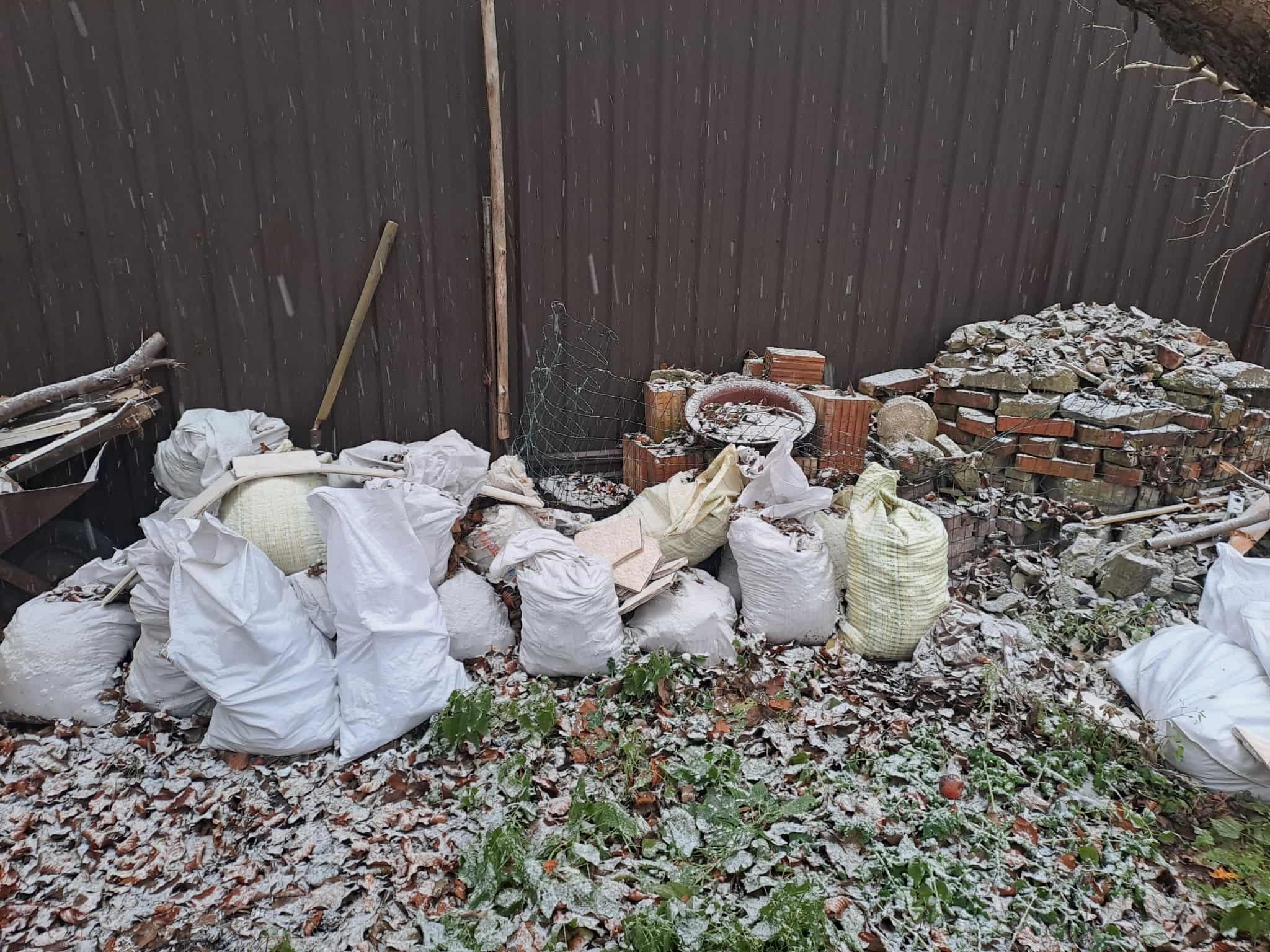 Sterta gruzu i worków z odpadami budowlanymi pod brązowym płotem, lekko przyprószona śniegiem. Widoczne cegły, potłuczone płytki i resztki roślinności.