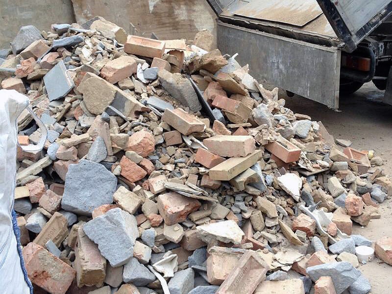 Stos gruzu z cegieł i betonu na ziemi, z otwartą skrzynią ciężarówki w tle. Widoczne fragmenty cegieł z napisem 'LONDON'.