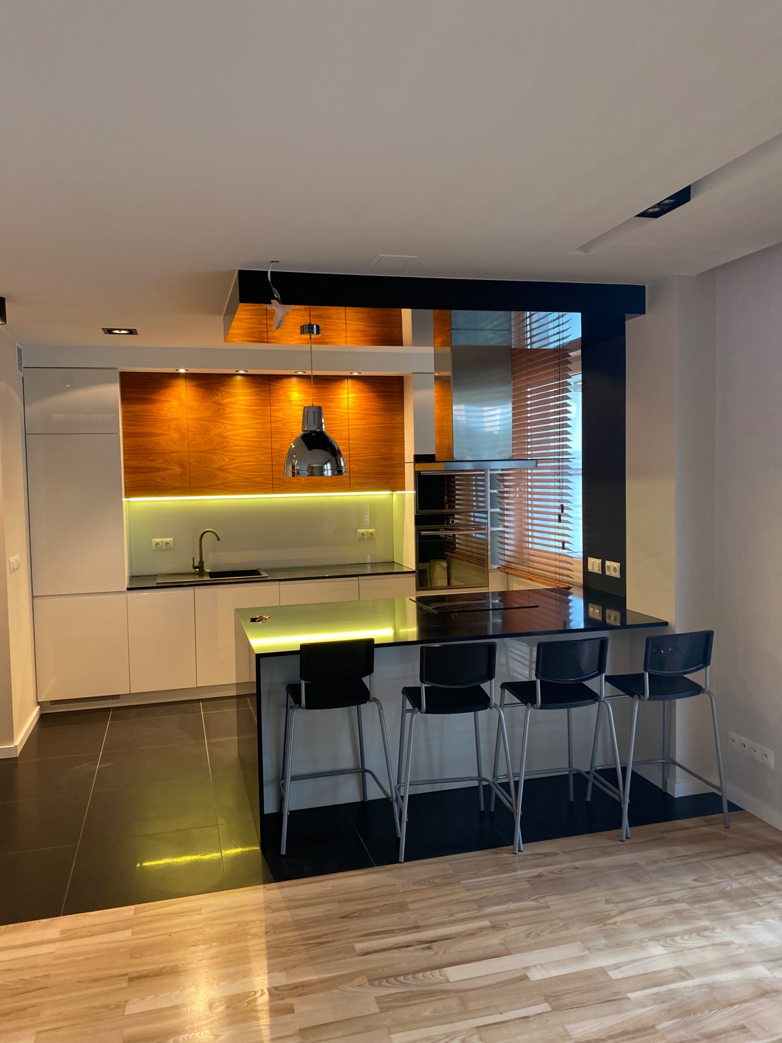 Nowoczesna kuchnia z wyspą i drewnianymi akcentami. Białe szafki kontrastują z ciemnym blatem i podłogą. Nad wyspą wisi designerska lampa. Widoczne drewniane żaluzje w oknie.